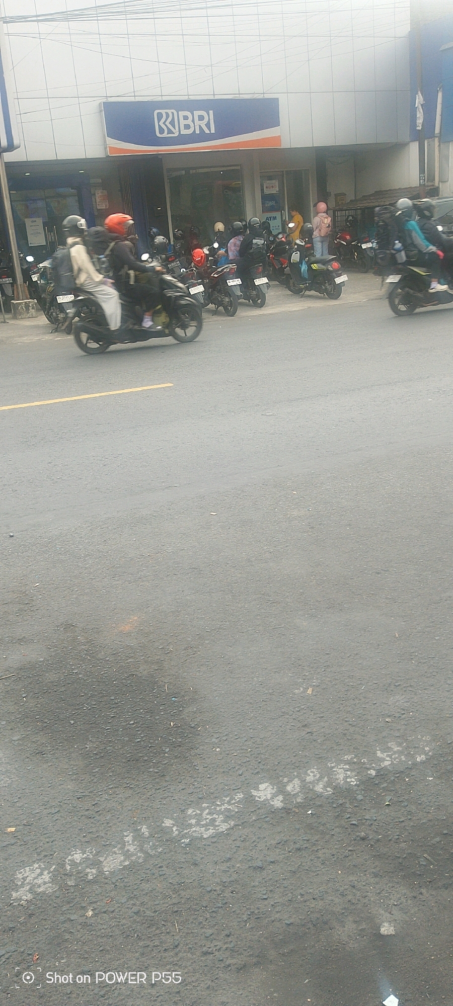 In an urban setting, possibly in the city of Unknown, Unknown, a bank with a prominent "BRI" logo on its facade is visible. The bank has glass doors and windows, with an ATM visible to the right of the entrance. A street with asphalt pavement runs in front of the bank. A yellow line is painted on the road, indicating a lane divider. A significant number of motorcycles are parked in front of the bank, suggesting it is a popular location. Several individuals are seen on or near motorcycles, some wearing helmets. One person is riding a motorcycle in the foreground, with motion blur indicating movement. The sky is overcast, suggesting a cloudy day. The overall scene depicts a busy commercial area with a focus on transportation by motorcycle.