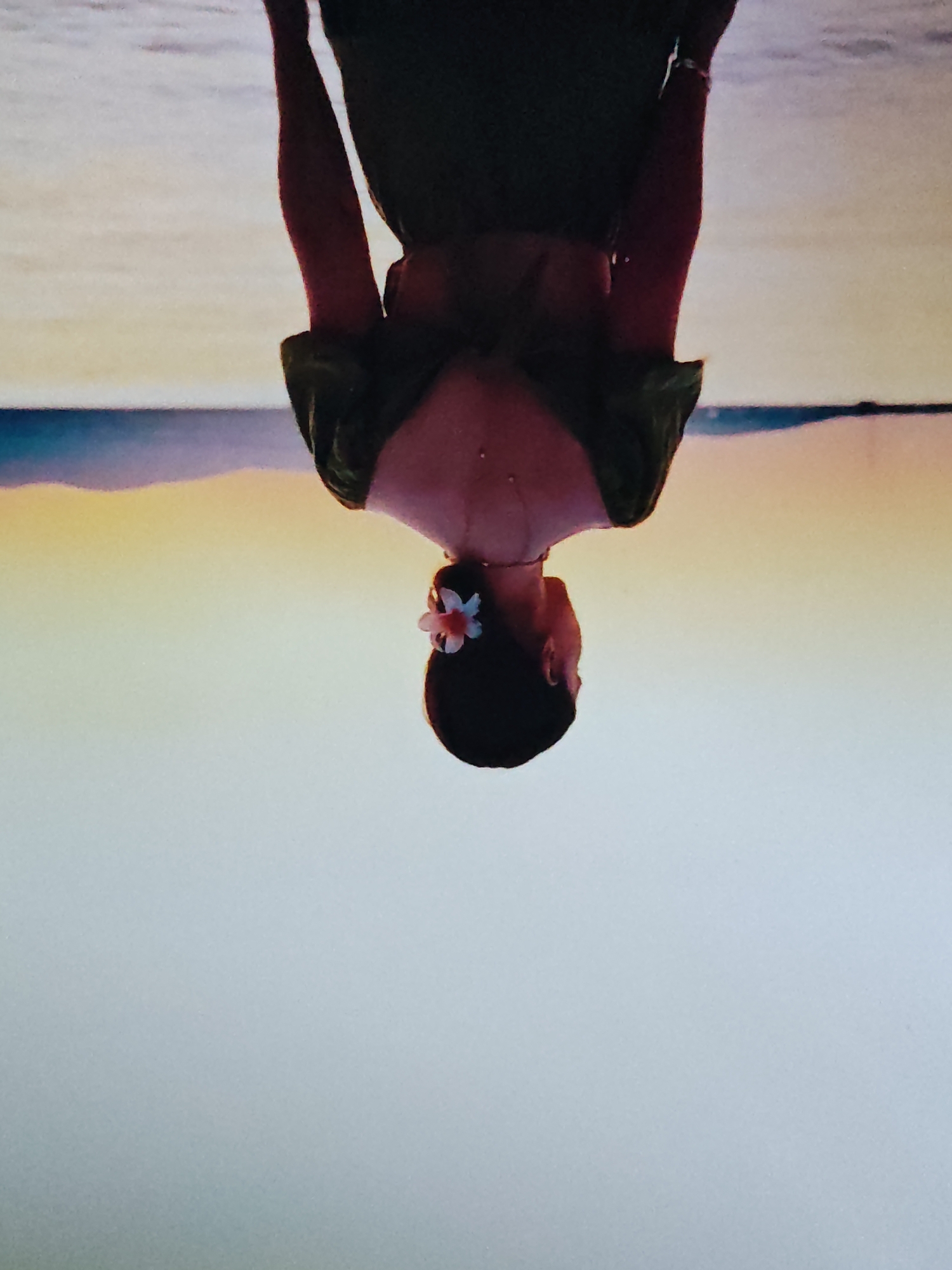 An upside-down medium shot captures a woman from the chest up, standing in a body of water at the edge of a beach. The woman, facing away from the viewer, has her arms raised and clasped behind her head. She wears a dark green, off-the-shoulder dress that drapes over her arms. A small, light pink flower is tucked into her dark hair, which is pulled back into a bun. Her back and shoulders are glistening with water droplets. The background consists of a calm sea or lake reflecting a gradient sky of pale yellow and light blue. The horizon line is visible in the distance, separating the sky from the water. The overall scene is serene and evokes a sense of peace. The location is described as Unknown, Unknown.