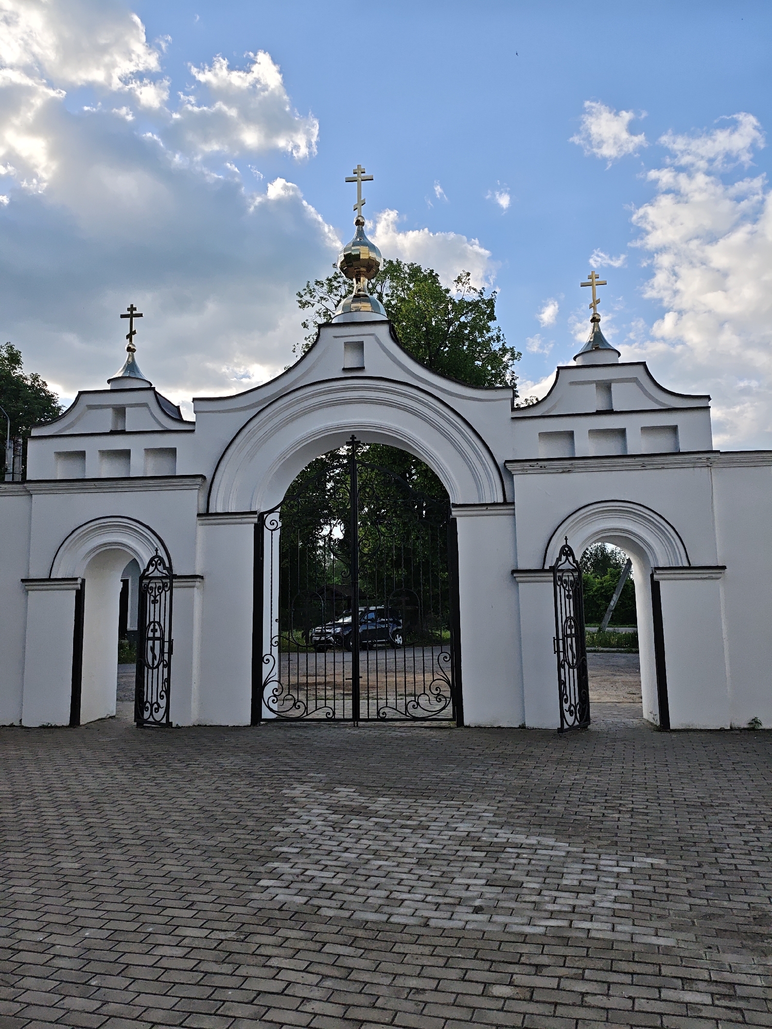 The image depicts the entrance gate to a religious building, likely a church or monastery, located in Боровково, Russia. The gate is painted white and features three arched openings: a large central arch with a black metal gate and two smaller side arches, each with a smaller black metal gate. Above each archway are decorative white structures topped with a cross, with the central structure also featuring a golden dome. The ground in front of the gate is paved with grey rectangular bricks. In the background, visible through the gate, are trees and a parked car. The sky is blue with scattered white clouds.