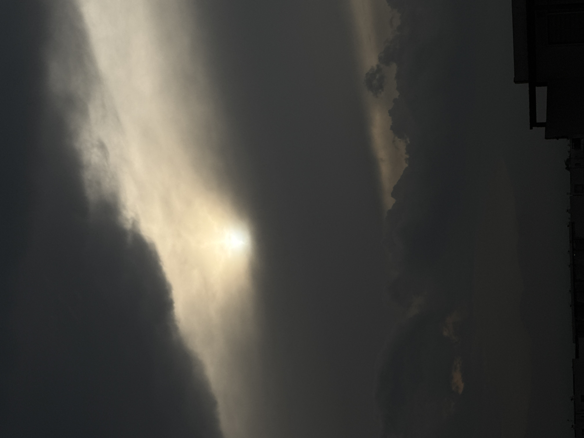 A low-angle, vertical shot captures a dimly lit sky. The sun, reduced to a bright, hazy orb, struggles to penetrate thick, dark clouds. Some building structures are visible along the right edge of the picture. The image seems to be from San Francisco Ocotlán, Ocotlán, Mexico.