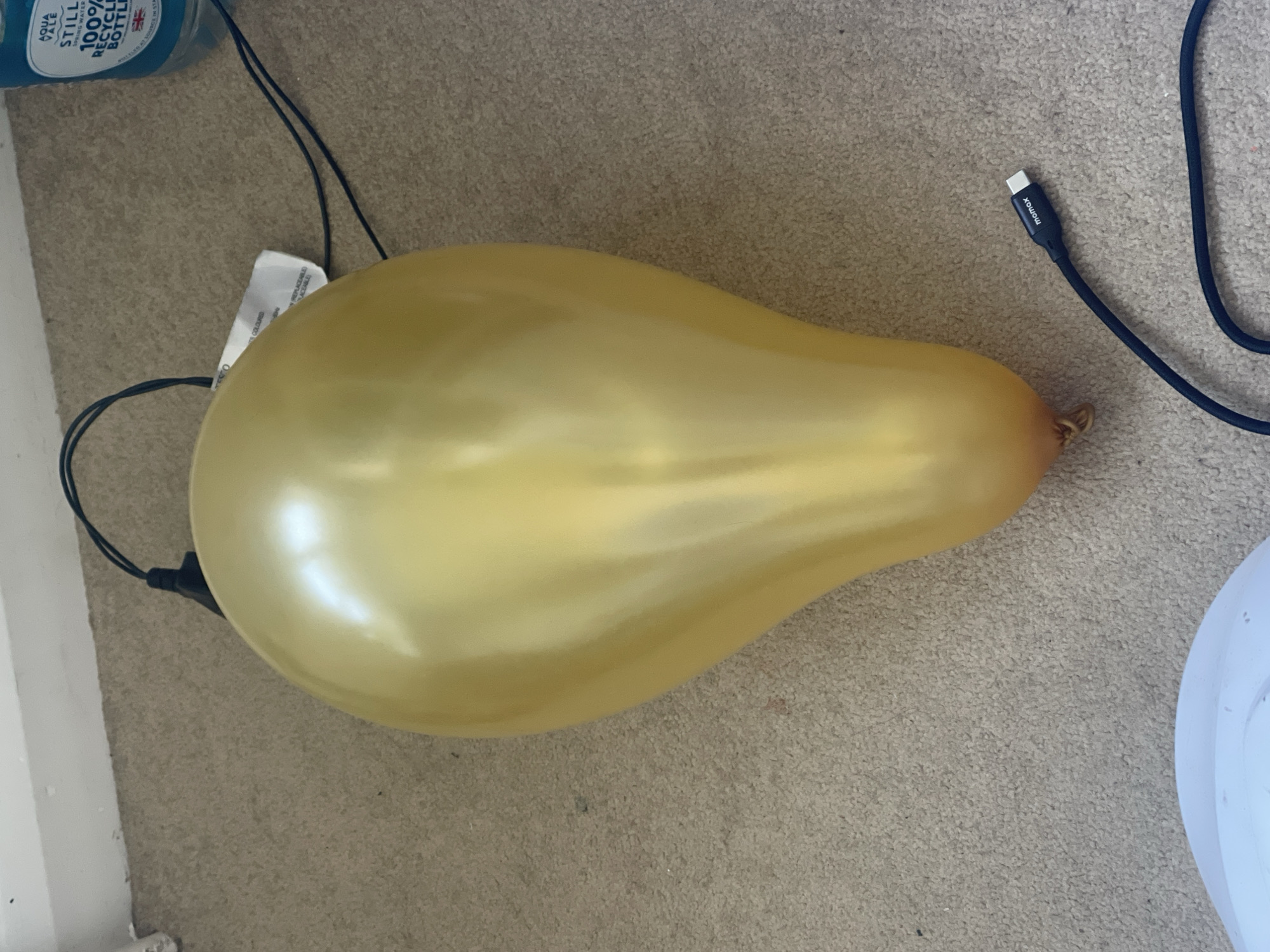 A gold pear-shaped balloon rests on a tan carpeted surface. Adjacent to the balloon are a blue water bottle labeled "Aqua Pale Still Spring Water 100% Recycled Bottle" and a black charging cable marked "momox". A white tag reading "TS Coloured" is partly visible near the balloon. The location of this image is in the city of Maidstone, United Kingdom.