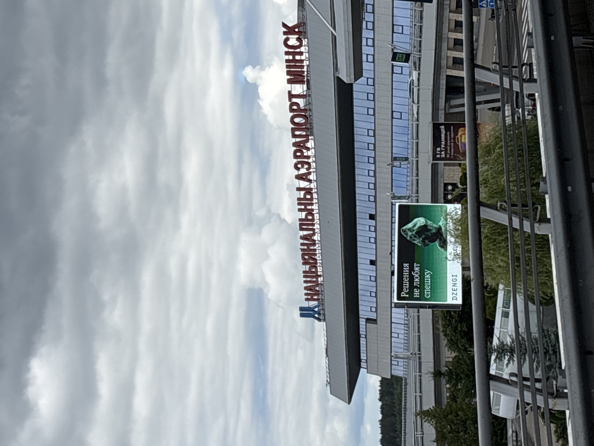 The image shows the National Airport Minsk, located in Belarus, under a cloudy sky. The airport building features large windows and is topped with the airport's name in red Cyrillic lettering. A large digital billboard displays an advertisement in Russian, featuring text that translates to "Decisions don't like haste" and an image of a green, rocky object. Another smaller advertisement board is visible to the right, with Russian text that can be translated to "5 GB abroad" and an image of a suitcase. In the foreground, a metal railing and a bridge structure obscure a portion of the view of the airport. Trees and other airport infrastructure are visible behind the railing.