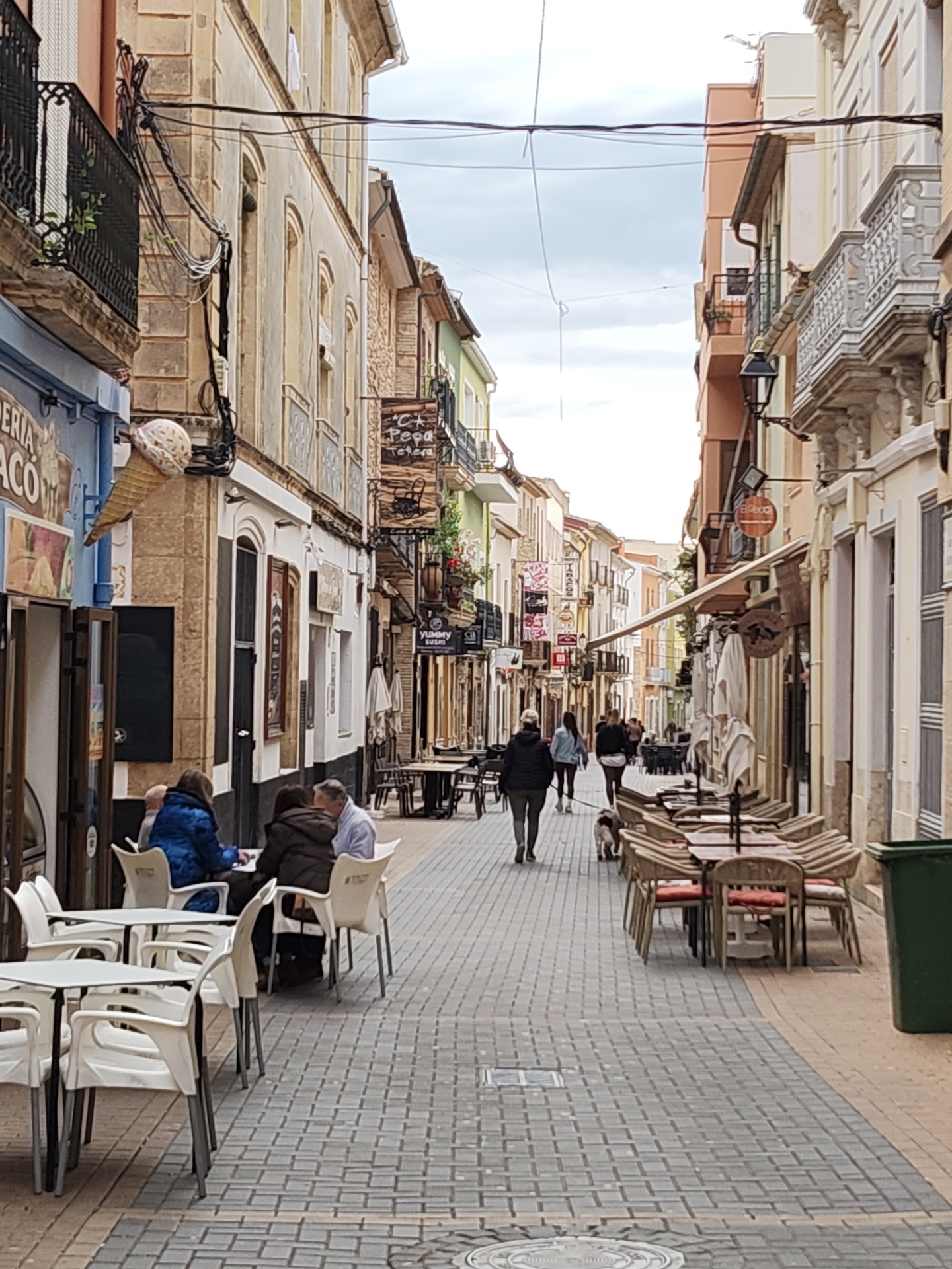 An eye-level, full outdoor shot depicts a street scene in the city of Dénia, Spain. The perspective is from the end of a narrow street that is flanked by buildings and various businesses. The street is paved with grey cobblestones.
On the left side of the street, four individuals are seated at a white plastic table outside of a restaurant. Further down the street are various businesses with signs, tables, and chairs set up outside. On the right side of the street, several tables and chairs are neatly arranged outside another establishment. A green trash bin sits on the edge of the sidewalk.
Several people are walking down the middle of the street away from the viewer, accompanied by a dog. The buildings lining the street are of varying heights and colors, with balconies and intricate architectural details. Overhead, wires crisscross the sky between the buildings. The sky is overcast and grey.