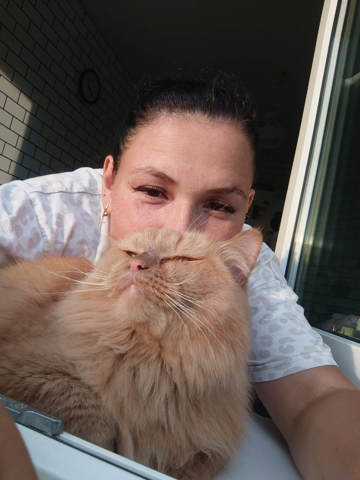 A selfie shows a woman with dark hair pulled back, leaning in towards the camera while holding a fluffy orange cat. The cat is positioned in front of the woman's face, obscuring her mouth and nose. The woman is wearing a white shirt with a light gray leopard print. In the background, a wall with white subway tiles is visible, along with a round clock. To the right, a window frame with white trim and reflective glass is present. The scene appears to be indoors, possibly in a home. The lighting suggests it is daytime, with sunlight casting shadows on the tiled wall. The image does not contain information to determine its location in Chaikovsky, Russia.