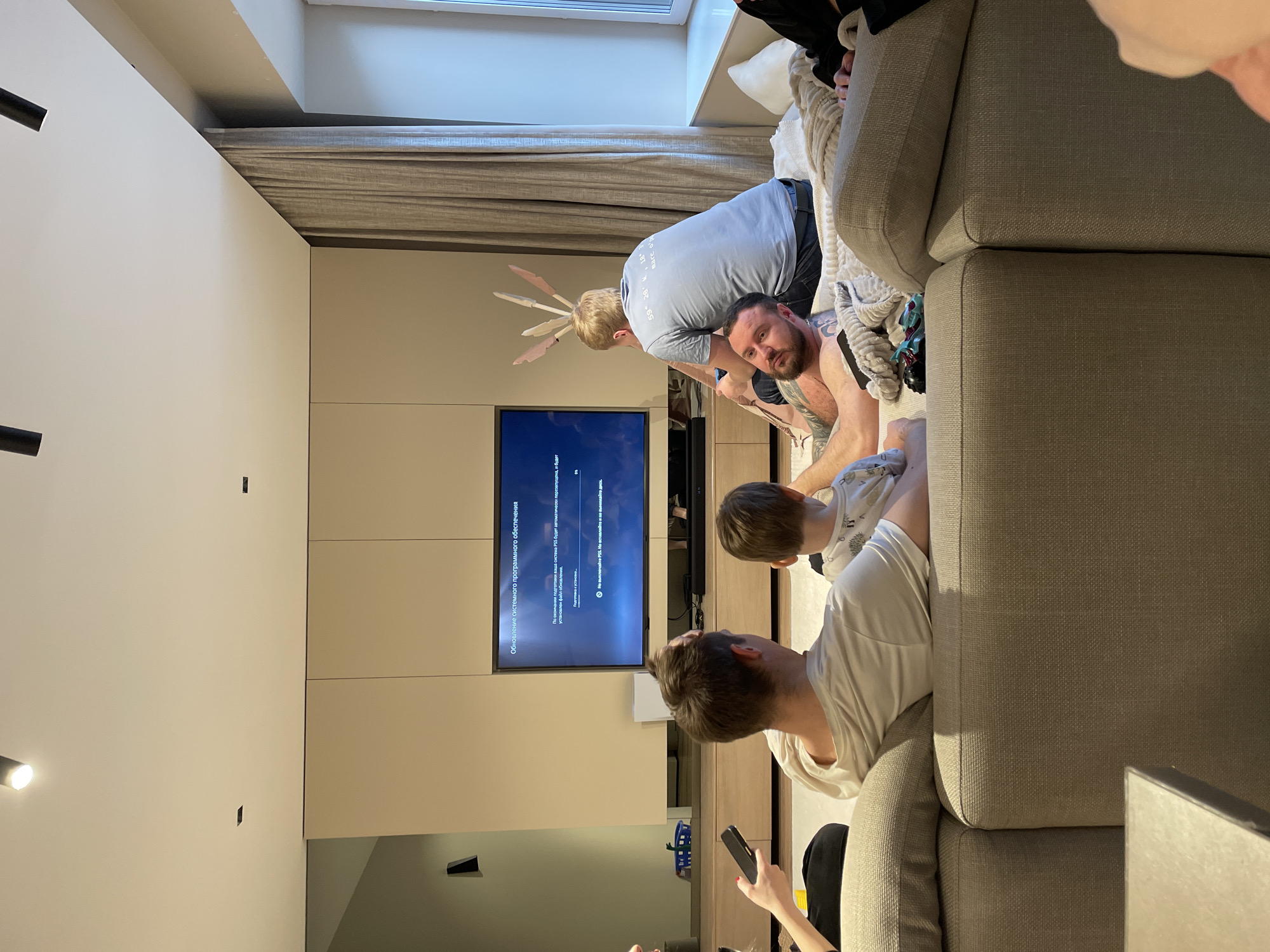 The scene shows a living room with four people: two adults and two children, one of whom is holding a phone. A man with bunny ears is leaning over another man. The children are closer to the sofa, looking at a TV screen which displays an update for a PlayStation 5 system. The room is decorated in neutral tones with a modern, minimalist style. It includes a large TV and a wide window. The OCR suggests the location might be associated with Krasnoyarsk, Russia.