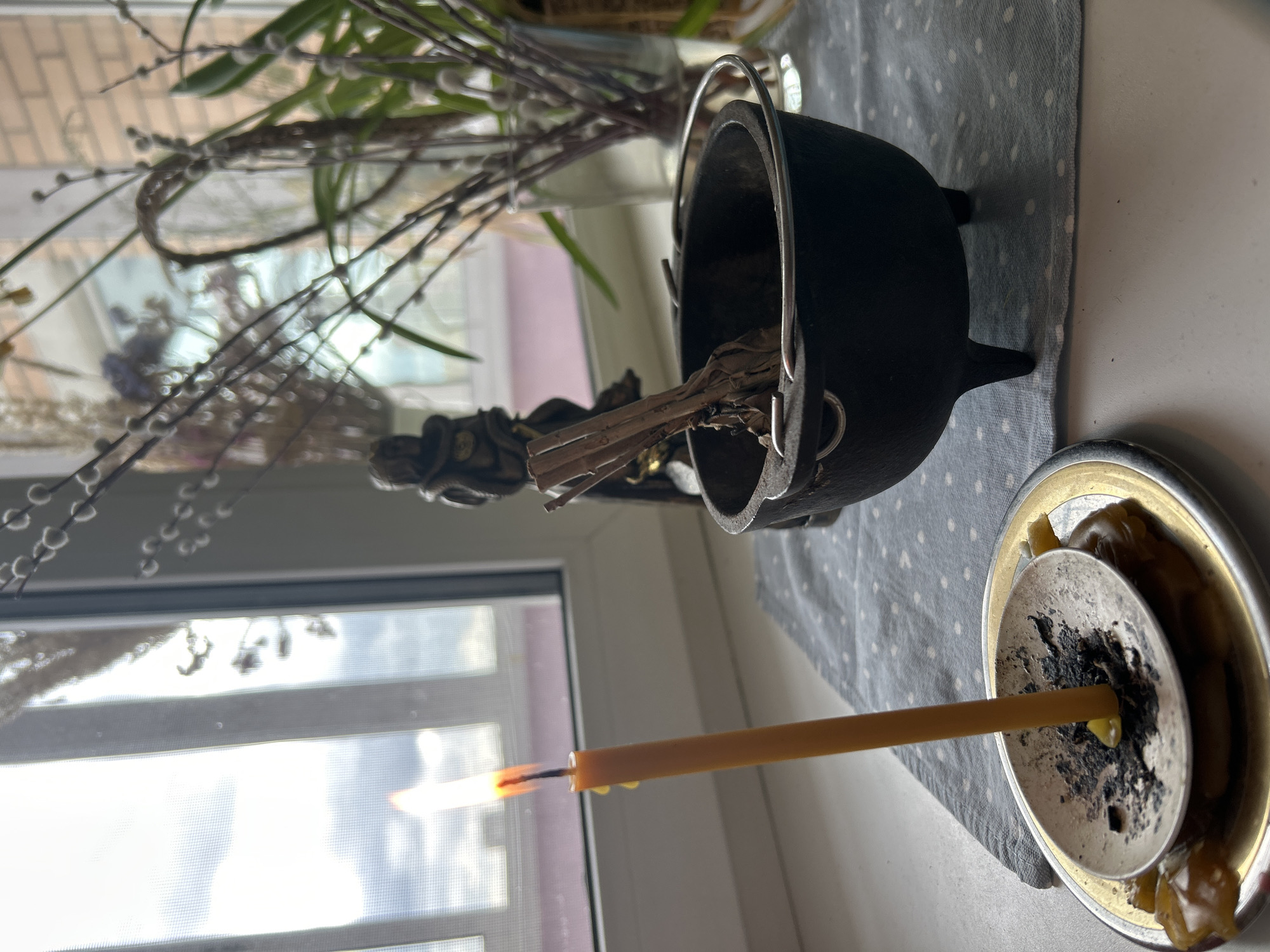 A windowsill scene includes a lit yellow candle in a metal candle holder with solidified wax and burnt wick residue, a black metal cauldron with a silver handle holding bundled sage, and a small statue in front of a window with pussy willows and other plants in a vase. The cauldron sits on a light grey cloth with white polka dots. The scene appears to be inside a residence. There is no confirmed evidence of the location being Balashikha, Russia.