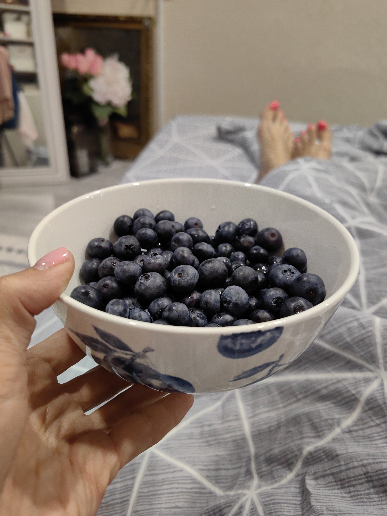 A close-up, eye-level shot shows a hand holding a white ceramic bowl filled with fresh blueberries. The bowl has a blue floral pattern on its exterior. In the background, out of focus, are a person's feet with pink nail polish, resting on a grey and white patterned duvet. Further in the background, a blurry image of a mirror with flowers inside it is visible. The scene appears to be indoors, possibly in a bedroom. There is no information that can help determine the location within the city of Fanipal, Belarus.