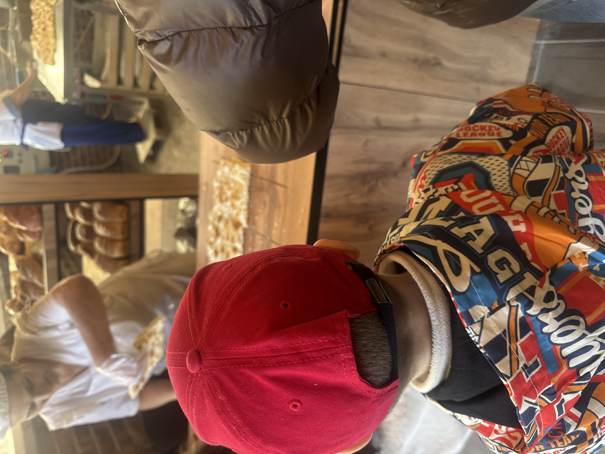 This image captures an indoor scene within what appears to be a bakery or food shop, set in Sochi, Russia. The primary subject is viewed from behind, featuring the back of a person's head and upper torso.
The person is wearing a bright red baseball cap with an adjustable strap visible at the back, covering dark hair. They are dressed in a colorful, patterned puffer-style jacket. The jacket's design is busy, featuring various graphics and text related to hockey, with "HOCKEY LEAGUE" clearly visible in multiple instances. Other words like "TEAM" are also present, alongside illustrations of hockey equipment such as sticks and pucks. A cream-colored, fleece-like collar or lining is visible beneath the jacket's main collar.
In the background, a display counter or shelf is laden with numerous baked goods, appearing to be bread or pastries, neatly arranged. Behind this counter, a second individual, likely an employee, is partially visible. This person is wearing a light-colored uniform, possibly a white top and blue bottoms, and appears to be tending to the products or serving customers. The environment suggests an active retail space, possibly bustling with daily operations. The lighting is bright and even, indicating daytime.