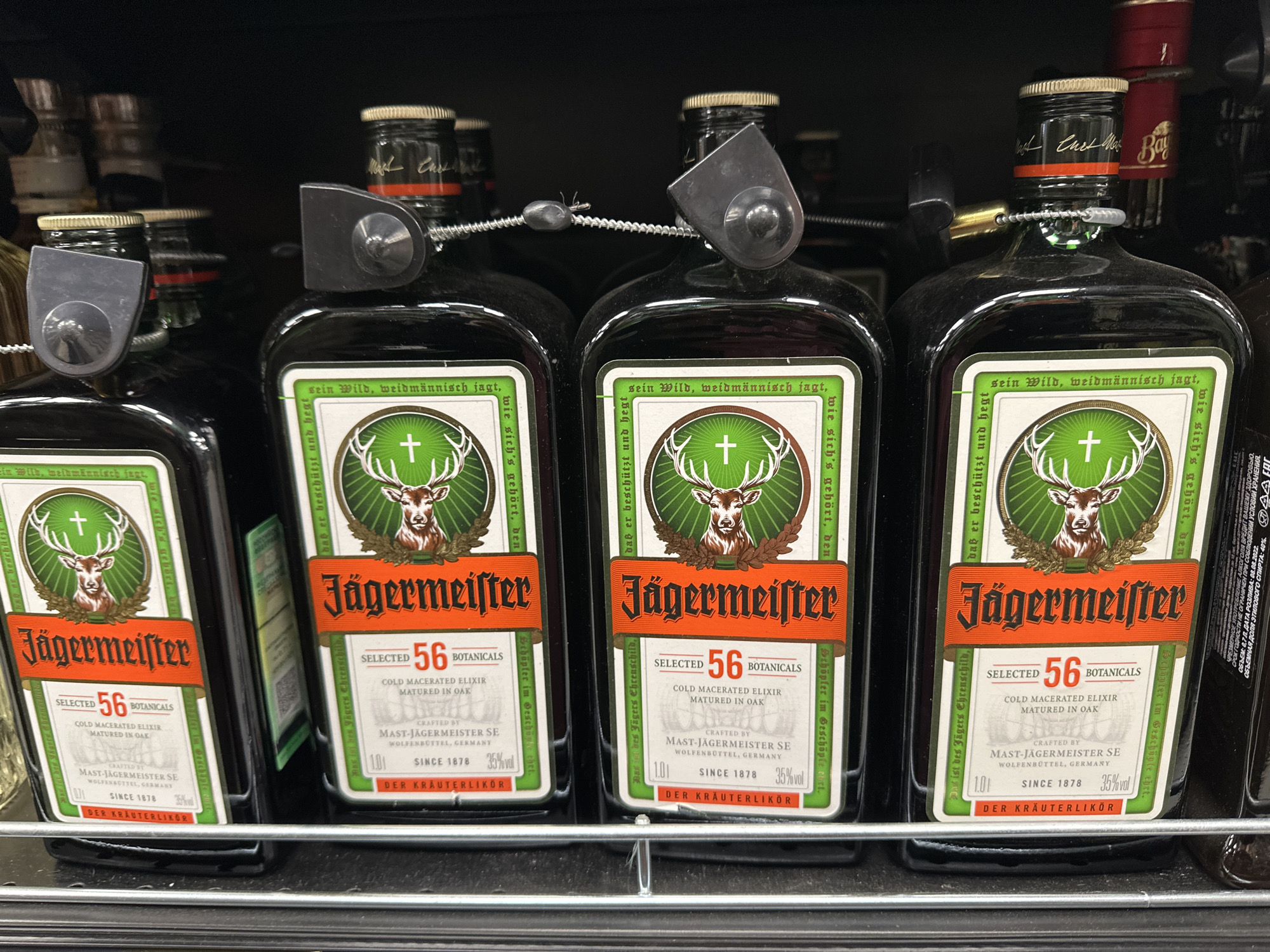 The image shows four bottles of Jägermeister liqueur on a metal shelf, likely in a store. Each bottle is fitted with a security device on its neck. The labels are clearly visible, displaying the Jägermeister logo with the deer and cross, the "56 Botanicals" claim, and other information in German. The bottles are aligned closely together, suggesting they are on a retail shelf. The presence of some Russian text on the right side indicates the location is likely Moscow, Russia.