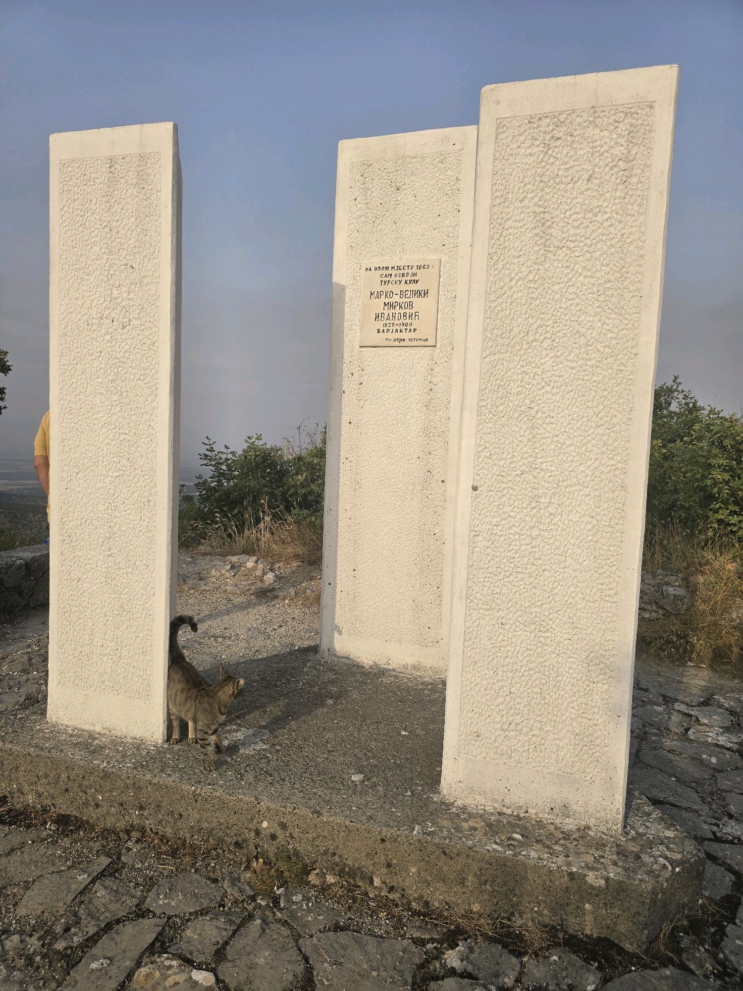 An outdoor scene features a tabby cat standing on a stone pathway in front of three large, rectangular, white concrete pillars. The cat is facing right, with its tail curved upwards. The middle pillar has a plaque with text in Cyrillic script. A person wearing a yellow shirt is partially visible to the left of the frame, standing behind the leftmost pillar. The background consists of dry grass, shrubs, and a hazy landscape under a pale blue sky. The ground is paved with irregular stones and gravel. The setting appears to be a historical monument site in Gornja Vrbica, Montenegro, possibly marking a significant location or event, given the plaque and memorial-like structure.
