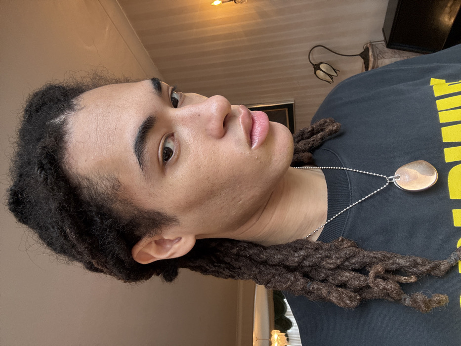 A close-up portrait depicts an individual with light-brown skin and dark dreadlocks, looking directly at the camera. The person is wearing a dark-colored top and a silver-colored ball chain necklace with an oval, two-toned pendant. The background is an indoor setting, likely a private residence or establishment. On the right, a light-brown wall with vertical striping is visible, featuring a wall-mounted sconce with a glowing, candle-like light source. Below the sconce, a dark piece of furniture and a decorative lamp with a curved stem and two inverted, tulip-shaped shades are present. A chandelier is partially visible in the upper left background. The specific location within London, United Kingdom, is not discernible from the visual evidence.