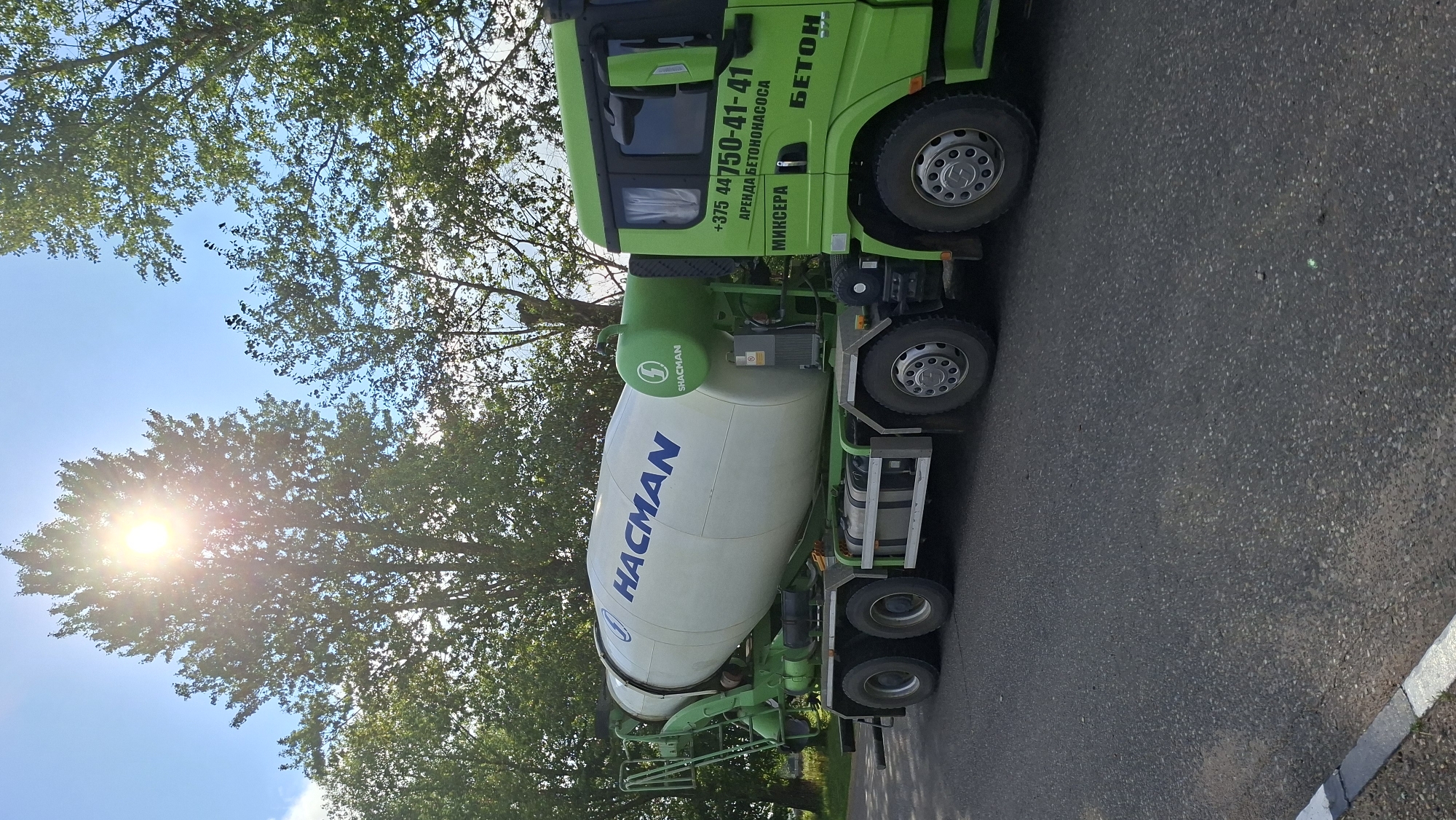 A bright, sunny day is captured in this image showing a green cement mixer truck parked on an asphalt road. The truck features a large white drum with the blue lettering "HACMAN" and the "SHACMAN" logo. The side of the truck's cab displays contact information: "+375 44 750-41-41", "АРЕНДА БЕТОНОНАСОСА", "МИКСЕРА", and "БЕТОН". The truck is positioned with its front facing right, and the drum is angled slightly upwards. Lush green trees line the background, and the sun is prominently visible, peeking through the leaves of a tall tree on the left, creating lens flare. The scene appears to be in a rural or suburban setting, possibly on the outskirts of Забалоцкі сельскі Савет, Belarus.