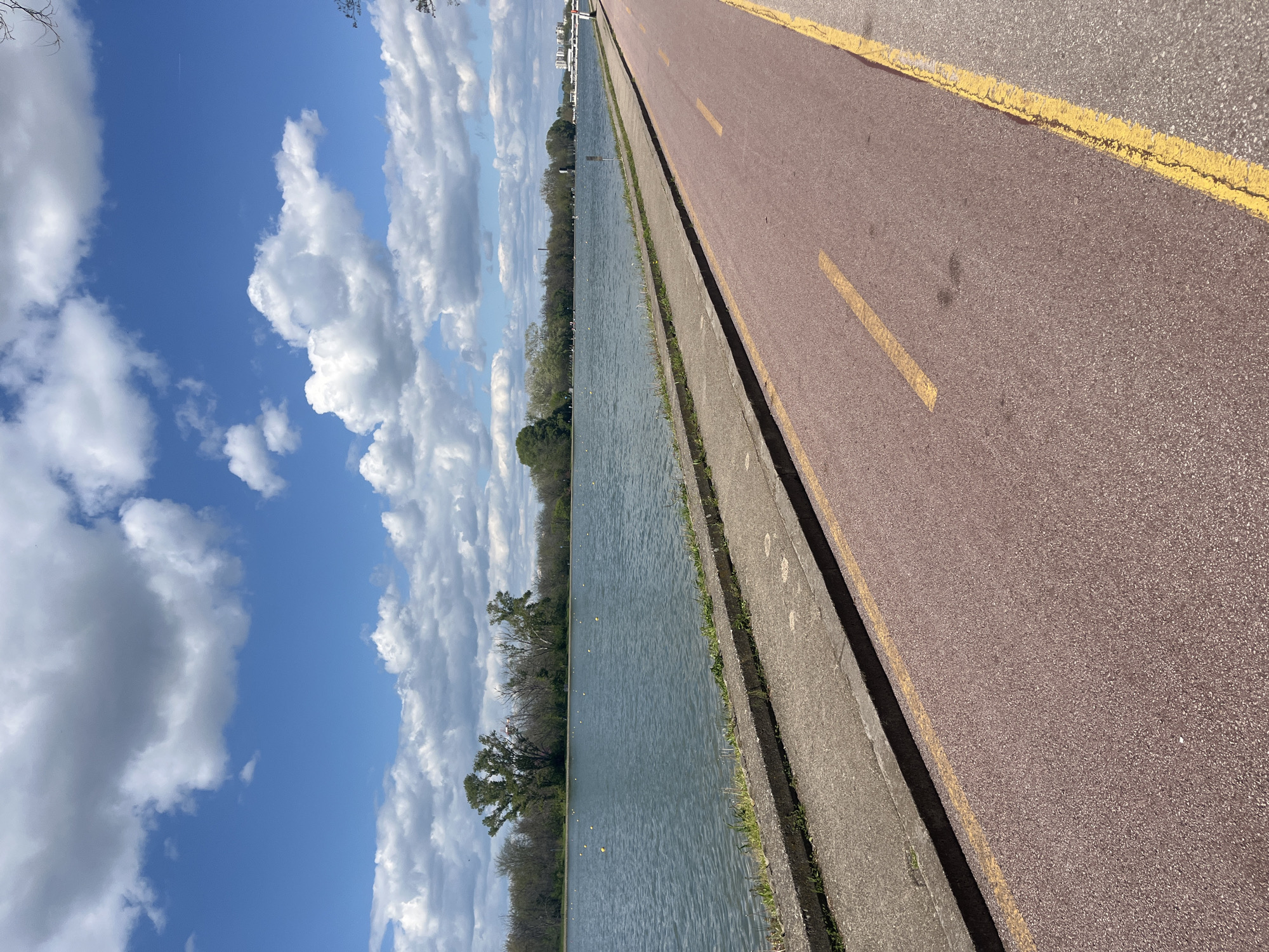 The image depicts a panoramic view of a long, straight body of water, likely a rowing canal or regatta course, alongside a paved pathway under a clear blue sky. The scene is oriented horizontally, showcasing a sunny day.
In the foreground and extending along the right side, a wide, reddish-brown asphalt path or road is visible, marked with distinct yellow lines that delineate lanes or edges. This path is separated from the water by a narrow concrete curb. The water itself appears calm and blue-grey, with small yellow buoys sparsely floating across its surface, suggesting a marked course for activities such as rowing.
On the far bank of the water, a dense line of trees, mostly deciduous with some bare branches and others showing early spring foliage, stretches into the distance. Beyond these trees, on the horizon, several modern buildings are discernible, indicating the proximity of a city. The sky above is a vibrant blue, filled with scattered, fluffy white cumulus clouds. The bright daylight creates clear visibility and a sense of openness.
The setting strongly resembles the Plovdiv Regatta Venue in Plovdiv, Bulgaria, known for its extensive rowing canal. No people or specific activities are visible, but the infrastructure suggests a recreational or sporting area. There is no visible text in the image.