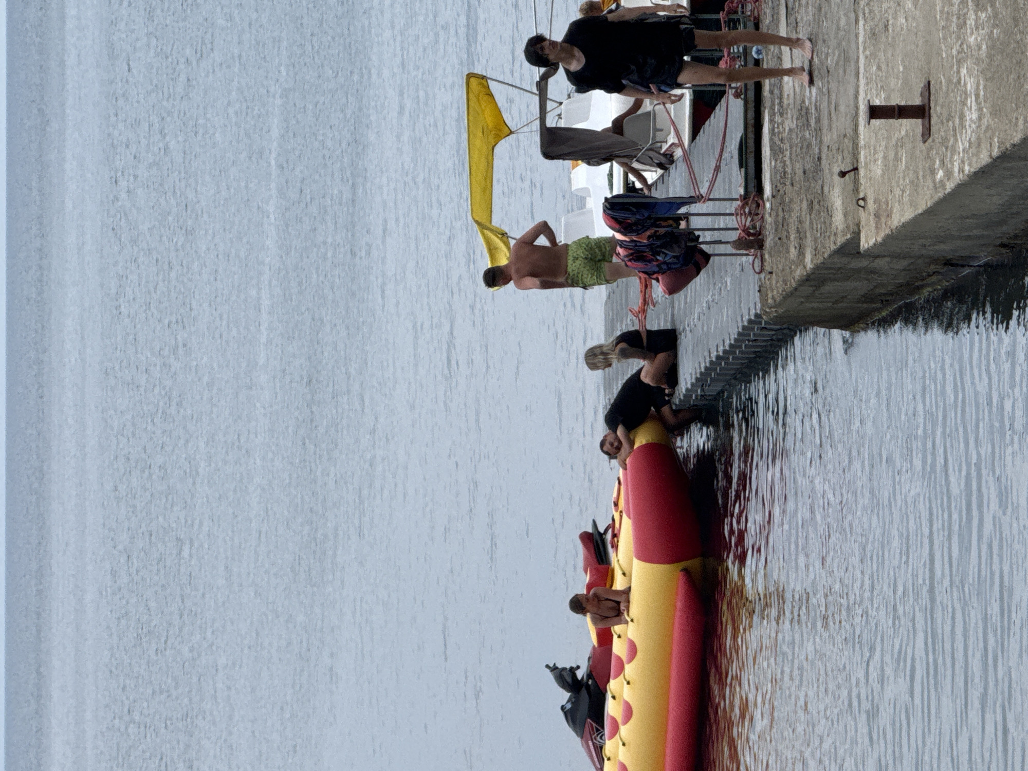 The image depicts a scene at a dock in Partenit, Ukraine. In the foreground, a concrete dock juts out into calm, grey water. Several people are present on and around the dock.
On the dock, to the left, a yellow and red inflatable banana boat is partially submerged in the water. A jet ski is attached to the back of the banana boat. A woman in a bikini is sitting on the banana boat. Further down the dock, another person is lying on the banana boat. To the right of the banana boat, a man in green shorts is standing, facing away from the camera.
Further along the dock, a person in a black t-shirt and shorts stands on the concrete, looking to the right. Several boats with yellow canopies are docked behind him. Life jackets and ropes are visible on the dock.
The background consists of a vast expanse of calm, grey water meeting a pale, overcast sky. The overall atmosphere is one of leisure and activity by the water.