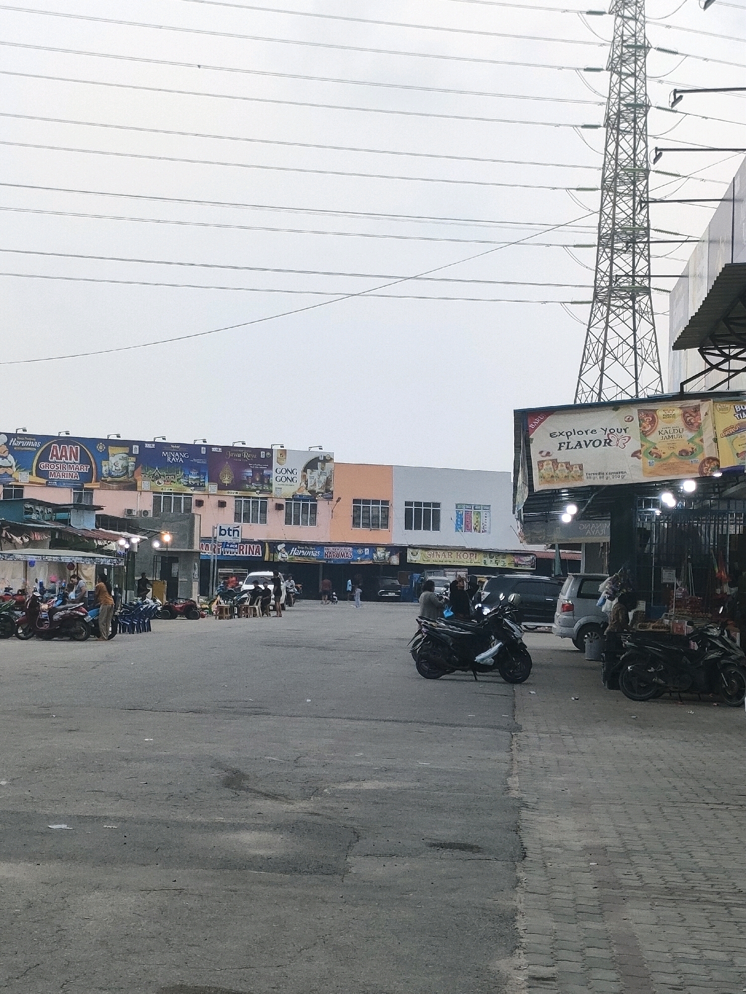 The image depicts an outdoor commercial area in Batam, Indonesia, possibly near a power transmission tower. The scene features a row of shops and stalls lining a paved lot. Several motorcycles are parked in the foreground and middle ground, some with riders. People are visible walking, interacting, and standing around. Overhead, power lines crisscross the sky, and a tall lattice transmission tower stands prominently on the right side of the frame. Billboards advertising various products, including food items like "Explore Your Flavor" and "Kaldu Jamur," are mounted on the buildings. The overall atmosphere suggests a busy marketplace or commercial hub.