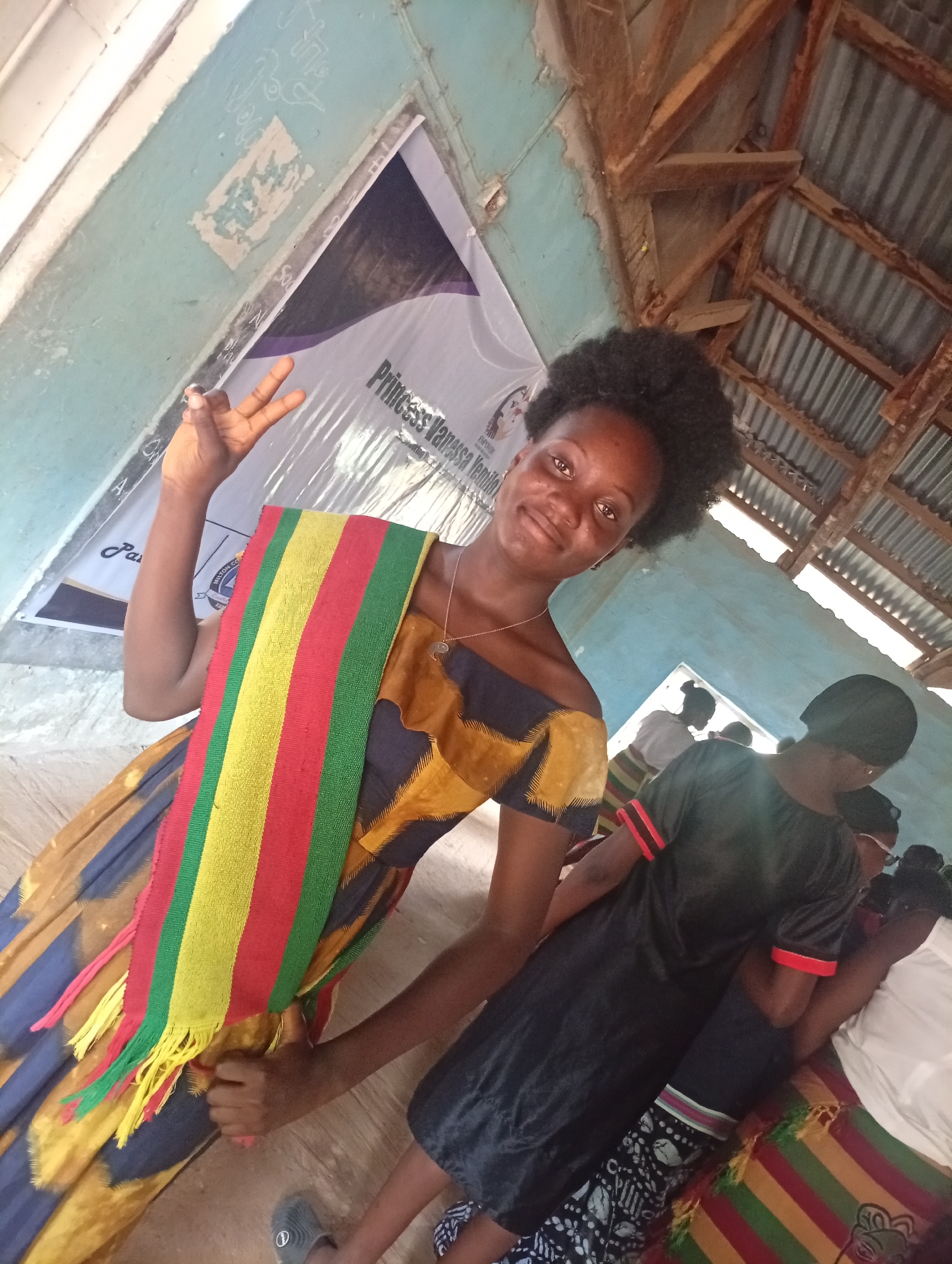 A young woman with dark, curly hair and brown eyes is posing for a photo in what appears to be an outdoor or semi-outdoor location. She is wearing an off-the-shoulder dress with a blue and yellow patterned fabric, and a colorful sash with red, yellow, and green stripes is draped across her body. Her right hand is raised, forming a peace sign with her index and middle fingers.
In the background, several other individuals are visible, some of whom are looking at cameras. A large banner with text that includes "Princess Vanessa Yenam" is partially visible behind the young woman. The environment suggests a community event or gathering, possibly in a place with rudimentary construction, as evidenced by the exposed wooden beams and corrugated metal roof. The walls are painted a light blue and show signs of wear. The ground appears to be dirt or concrete.