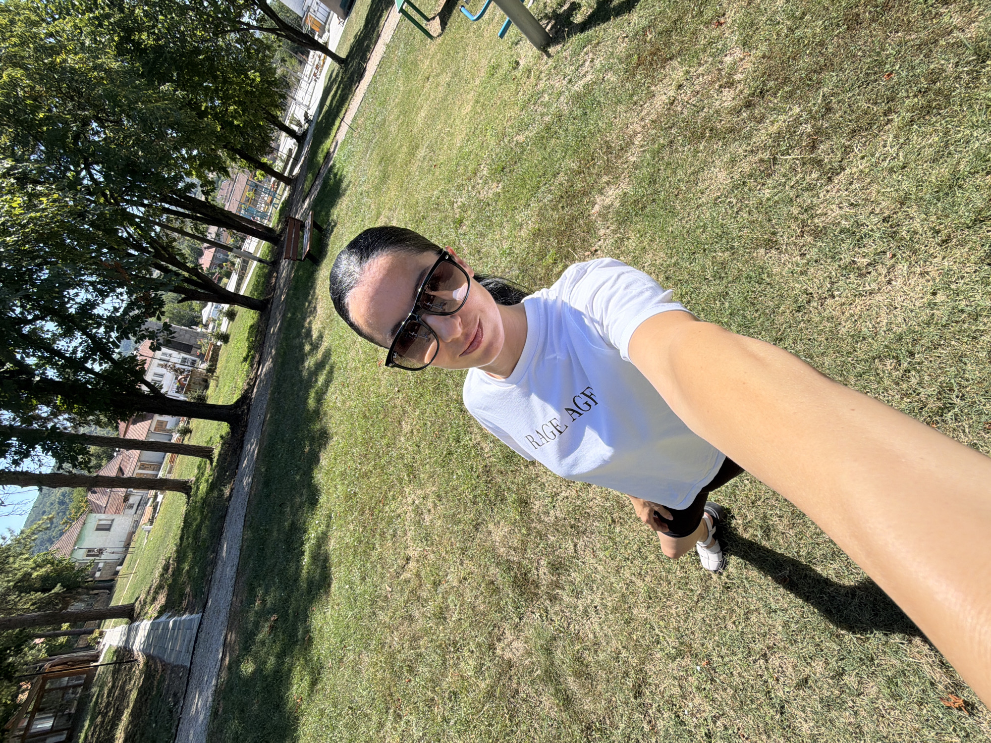 In the city of Chavdartsi, Bulgaria, on a sunny day, a woman stands in a grassy park. She is wearing a white t-shirt with "RAGE AGF" printed on it, black shorts, and white sneakers. Large, dark sunglasses obscure her eyes, and her dark hair is pulled back into a ponytail. She is holding a phone to take a selfie, her arm extending into the foreground. The park features a paved pathway, trees, benches, and children's playground equipment in the background. Residential buildings are visible behind the trees. The grass is somewhat dry, indicating a warm season.