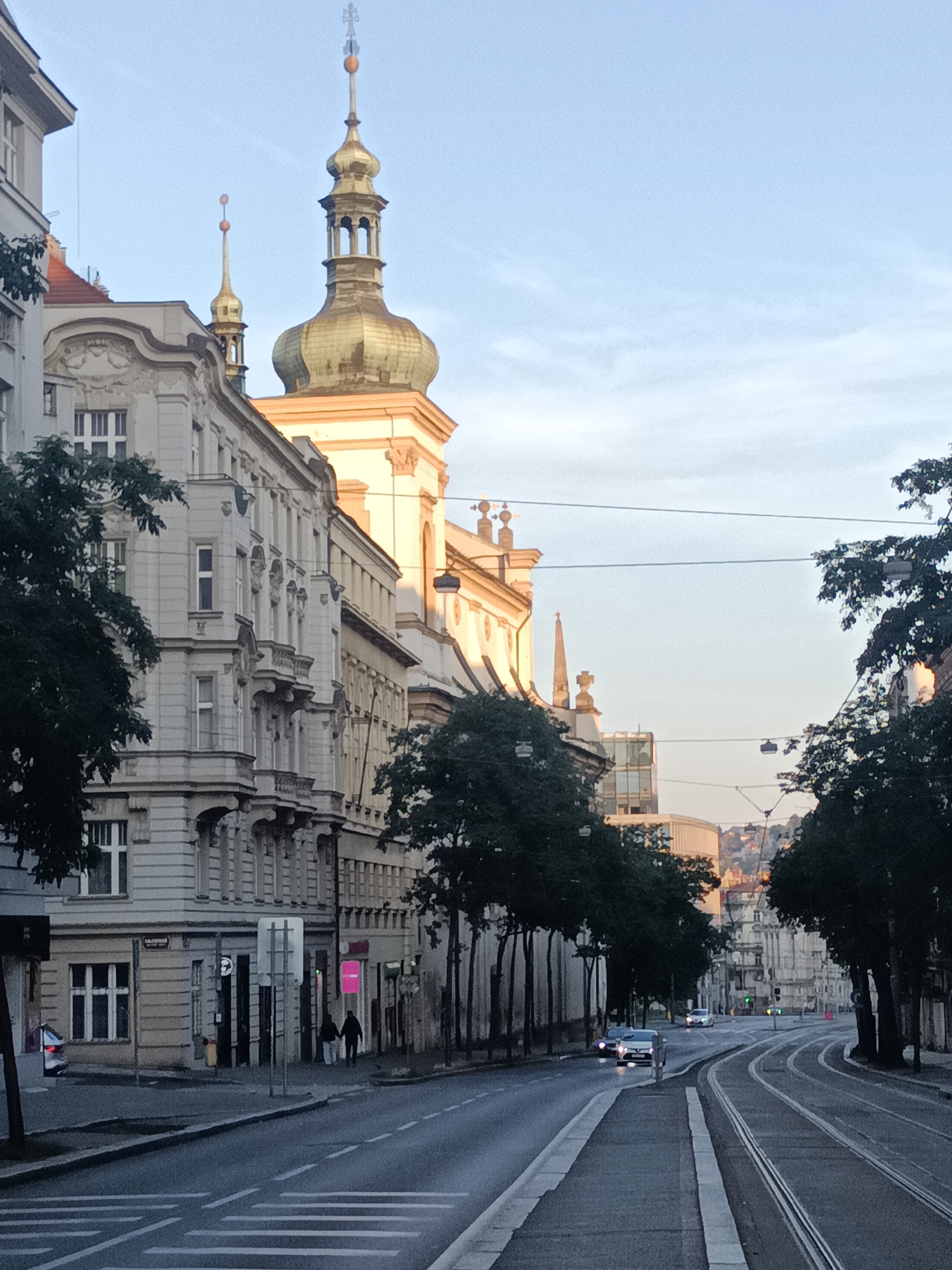 The image depicts a street scene in Prague, Czechia, likely in the Old Town or Malá Strana area, given the architectural style. The perspective is from the street level, looking down a road lined with ornate, historical buildings. The time of day appears to be early morning or late afternoon, as evidenced by the soft, warm light illuminating the facades of the buildings and the clear, pale blue sky.
Dominating the skyline on the left are two distinct golden spires of a church or cathedral, with intricate architectural details. The building directly in front of these spires is lighter in color and features multiple floors with numerous windows and balconies. Trees with dense green foliage are planted along the side of the road, casting shadows and framing the view.
The road itself is paved and features tram tracks running down the center. A pedestrian crossing with white stripes is visible in the foreground. There are several cars on the road, including a white car in the middle distance and another silver car closer to the foreground, both appearing to be in motion. Two figures, a man and a woman, are walking hand-in-hand on the sidewalk to the left, in front of a building with shop fronts.
Further down the street, the road curves, and more buildings are visible, some modern with glass facades, interspersed with older structures. The background shows a hazy cityscape with buildings ascending a hill. The overall atmosphere is peaceful and urban, capturing the architectural grandeur of Prague.