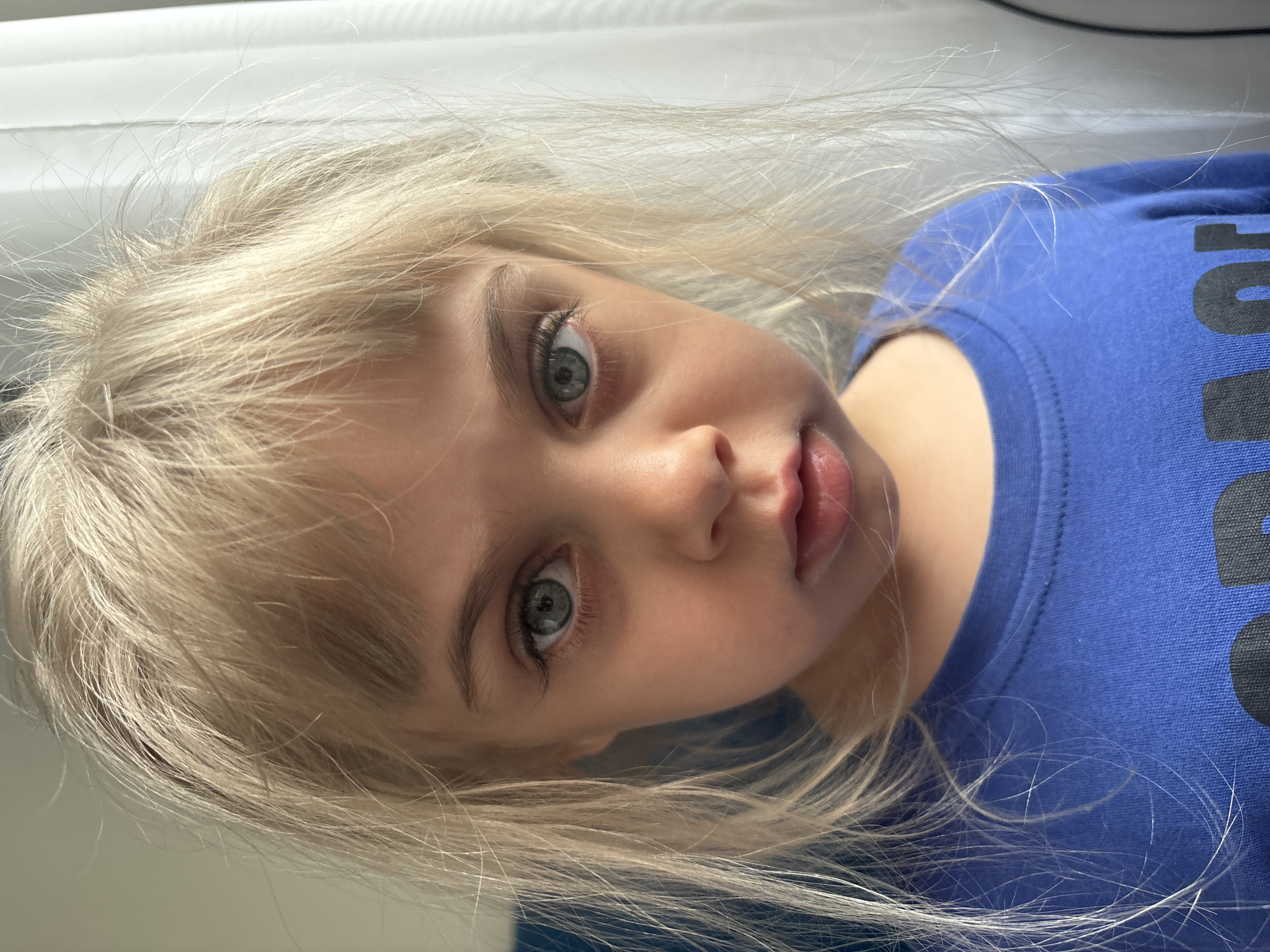 A close-up portrait shows a young child with pale blonde hair and pale skin. The child has light-colored eyes and is wearing a blue shirt. The child's gaze is directed towards the camera. The background is a white window. The image appears to be taken indoors. The location is Unknown, Unknown.
