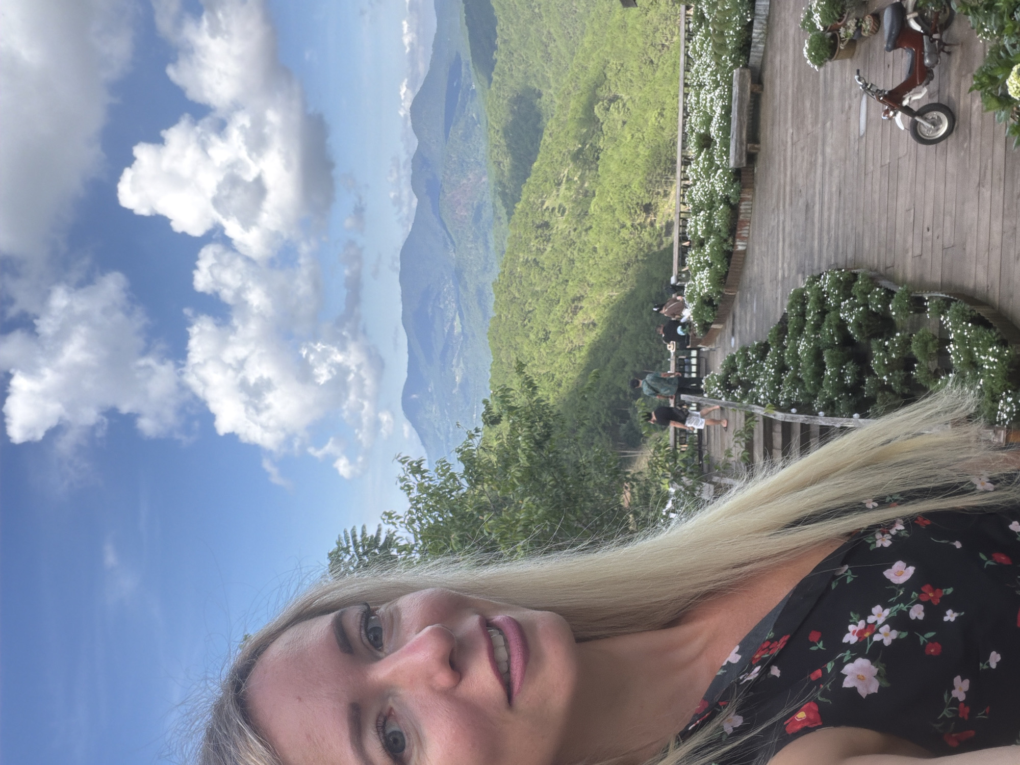 This is a selfie of a Caucasian woman with blonde hair and blue eyes, wearing a black floral dress. She is in Xuan Truong, Da Lat, Vietnam, with a scenic view of rolling green hills and mountains in the background under a partly cloudy blue sky. In the foreground, there is a wooden deck with several people walking and sitting. There are also planters filled with white flowers and a red and white moped parked on the deck.