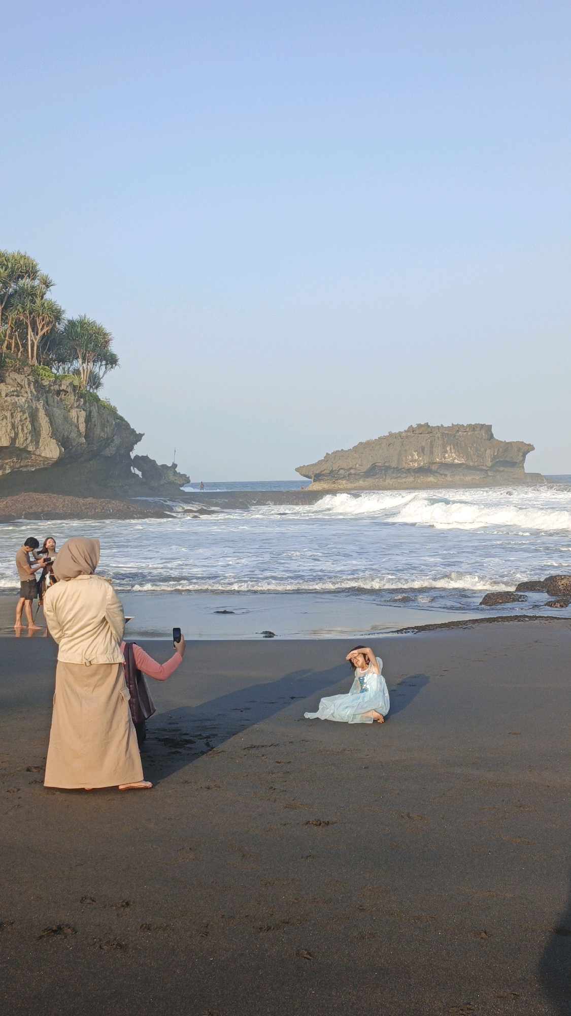 The image depicts a beach scene in Unknown, Indonesia, with a focus on a group of people interacting near the shoreline. In the foreground, a woman wearing a tan hijab and a long tan skirt is taking a photo with her phone. To her left, another person in a pink hijab is also holding up a phone, possibly taking a photo or recording. In the middle ground, a person in a light blue dress is sitting on the black sand, covering their eyes from the sun. Further in the background, a couple stands near the water's edge, with one person holding a camera. The environment includes a black sand beach, the ocean with moderate waves, and rocky outcrops with vegetation on the left. A large rock formation is visible in the middle of the ocean. The sky is clear and a pale blue. The overall lighting suggests it is late afternoon.