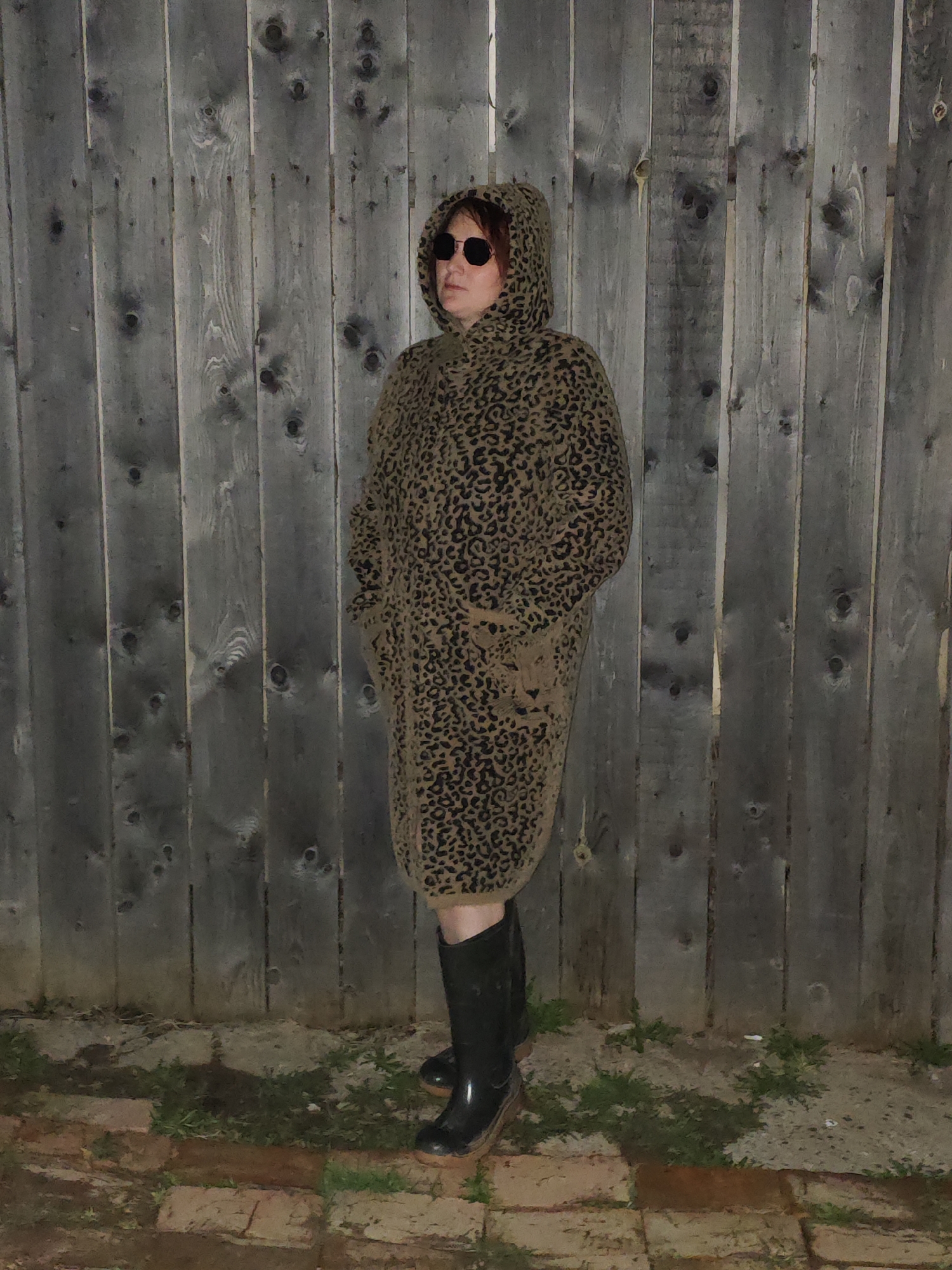 A person stands in front of a wooden fence, wearing a leopard print hooded coat and black rain boots. The person also wears sunglasses. The ground is brick and grass. The location is not specified in the image.