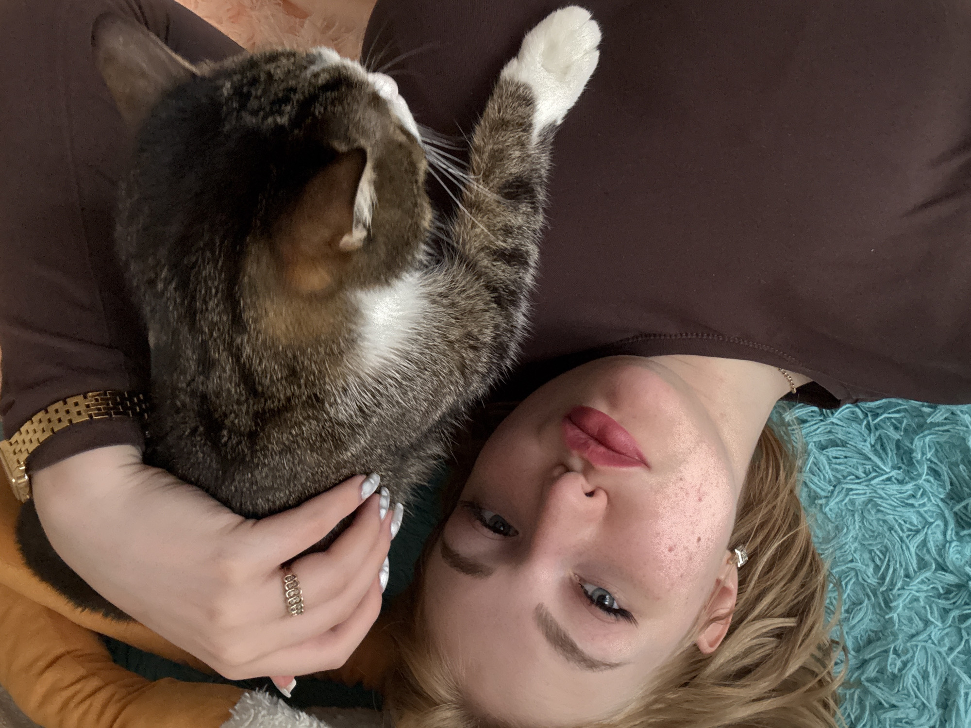 A young blonde woman lies on her back, her face turned towards the camera. She is wearing a brown shirt and makeup, with freckles visible on her skin. Her left hand, adorned with a ring and painted nails, rests on a tabby cat. The cat is lying on the woman's chest, its head turned away from the camera. A golden watch is visible on the woman's wrist. The background consists of a brown garment and a textured teal surface, possibly a blanket or rug. The overall impression is a close-up, intimate shot of the woman and her pet. The location is not specified in the image.
