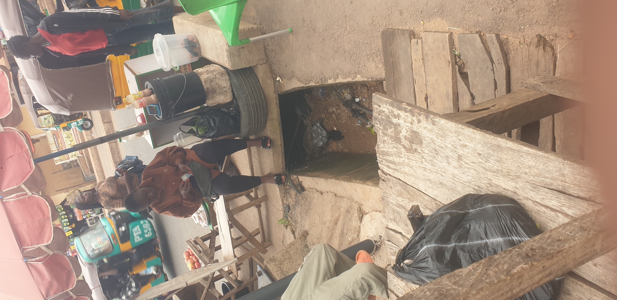 The image depicts a scene at a street market in Barakin Lerawi, Nigeria. Several people are visible, with one person in the foreground wearing a brown jacket and black leggings, holding money. Another person in the background is wearing a red and black jacket. There are various objects including bags, buckets, a tire, a green funnel, and a small vehicle resembling a tricycle, likely used for transport or vending.
The setting appears to be an outdoor market with dirt and concrete ground, and wooden planks forming a makeshift structure. A drain or open channel is visible, containing some debris. The overall atmosphere suggests a busy market day.
The lighting indicates it is daytime. There are no strong indicators of specific weather conditions, but the ground appears dry.
Visible text includes "PTE 6583" on the tricycle and some text on a sign in the background that is difficult to read completely but appears to include "LONE CUISINE". The image is oriented with the top edge on the left.