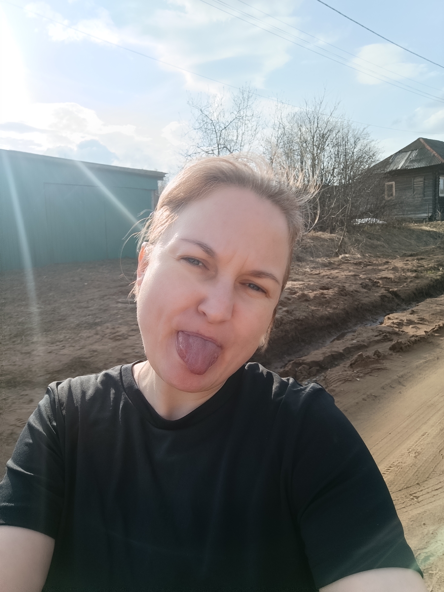 The image shows a fair-skinned woman with short blonde hair wearing a black t-shirt, sticking her tongue out at the camera. The background consists of a rural scene. There is a green metal building on the left, and a wooden house with a grey roof on the right. The ground is dirt. Bare trees are visible between the buildings. The sky is blue with some clouds. The location is Dubovaya, Russia.