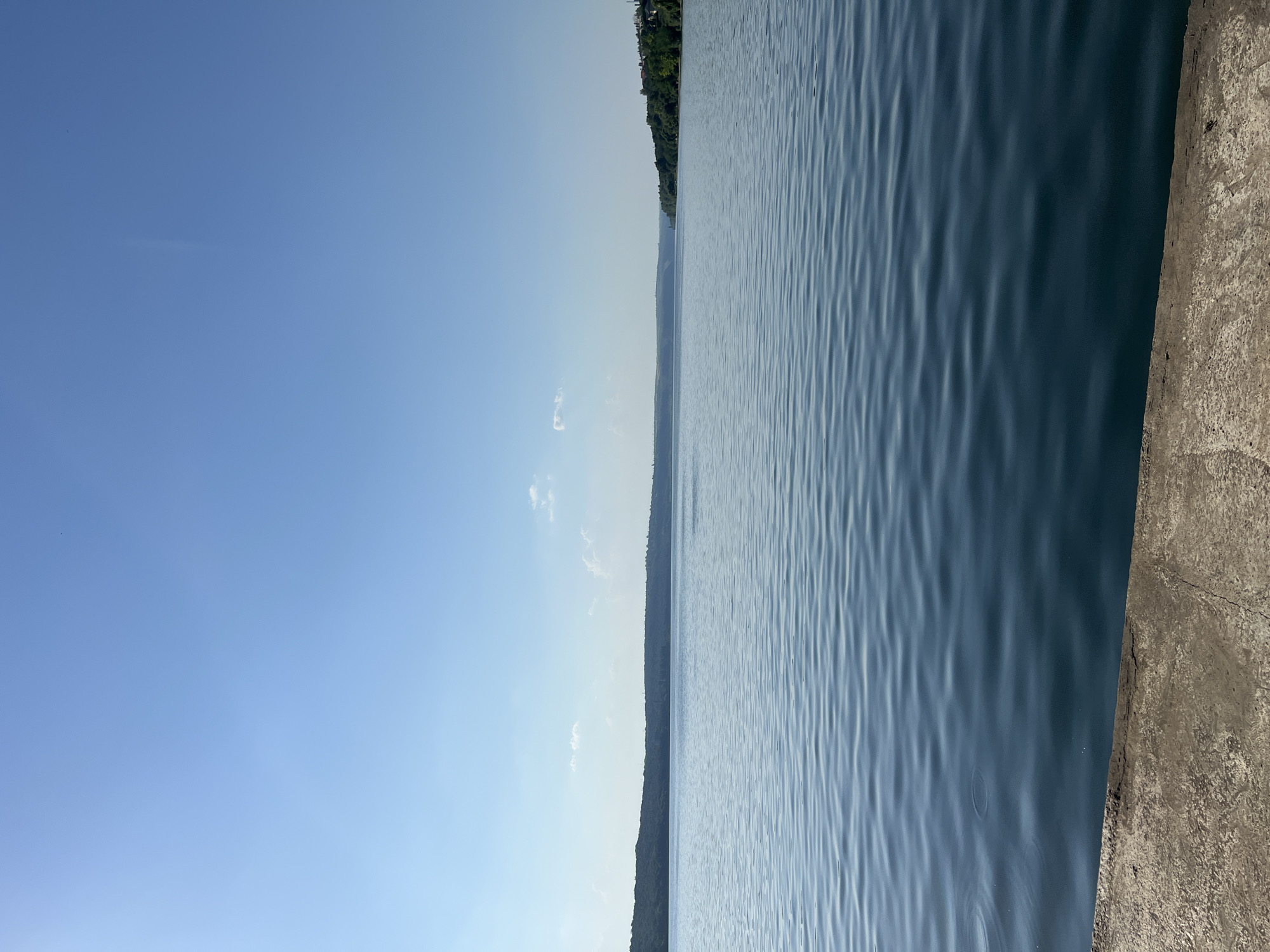 The image shows a wide, calm river with a clear blue sky. The foreground features a concrete embankment. In the distance, a treeline marks the opposite bank, with hints of buildings visible on the right side of the frame. The water has gentle ripples. The sky is mostly clear, with a few wispy clouds. The overall impression is one of a serene natural landscape, possibly within or near the city of Korzhevo, Moldova, given the contextual information.
