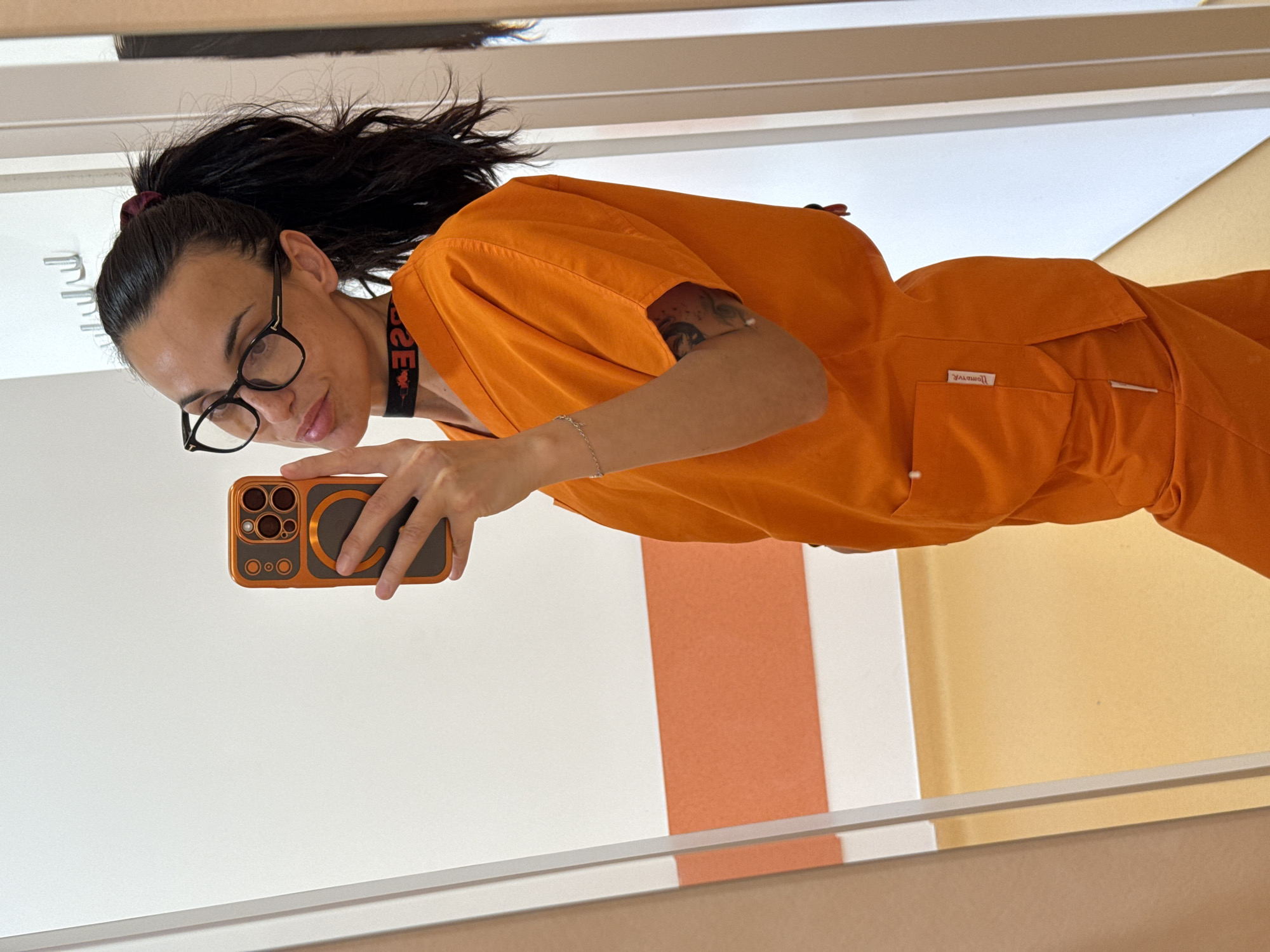 A woman with dark hair pulled back in a ponytail and wearing glasses is taking a selfie with her phone in a mirror. She is wearing an orange scrub suit. The background shows a white wall and a portion of an adjacent peach-colored wall. The location and city of Pleven, Bulgaria are not discernible from the image.