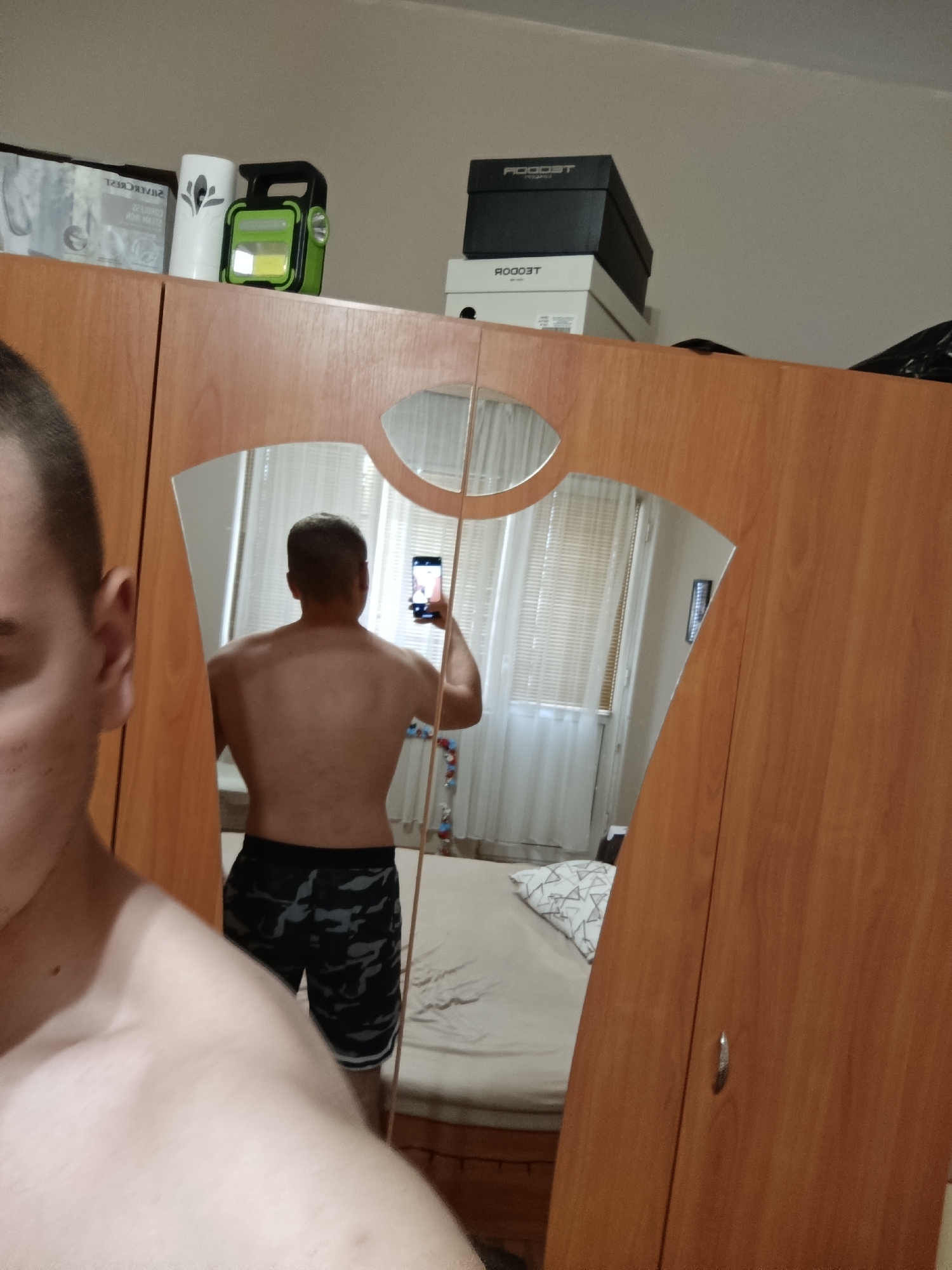 The image shows a mirror selfie taken by a person in what appears to be a bedroom in Rakovski, Bulgaria. In the foreground on the left, a portion of the person's shirtless left shoulder and the side of their head are visible. The main subject is seen in the reflection of a large, irregularly shaped mirror on a wooden wardrobe. The person, viewed from behind, displays their back and shoulders, holding a smartphone up to take the photograph. They are wearing dark-colored shorts with a camouflage pattern.
On top of the light brown wooden wardrobe, several objects are placed. To the left, there's a white cylindrical item, a green and black portable lantern or flashlight, and a white package showing the reversed text "SILVERCREST". To the right, a stack of shoe boxes is visible, with a black box on top and a white one underneath, both displaying the reversed brand name "ROOST".
The reflection reveals the interior of the room: a bed with light-colored bedding and a white pillow patterned with geometric shapes. Behind the bed, a window with closed Venetian blinds and white curtains is seen. A white wall extends to the right, where a vertical wall-mounted light fixture is visible. A colorful garland or decoration hangs near the window. The indoor setting appears to be illuminated by artificial light, implying it might be evening or night. The mirror's curved edges create slight distortions in the reflection.