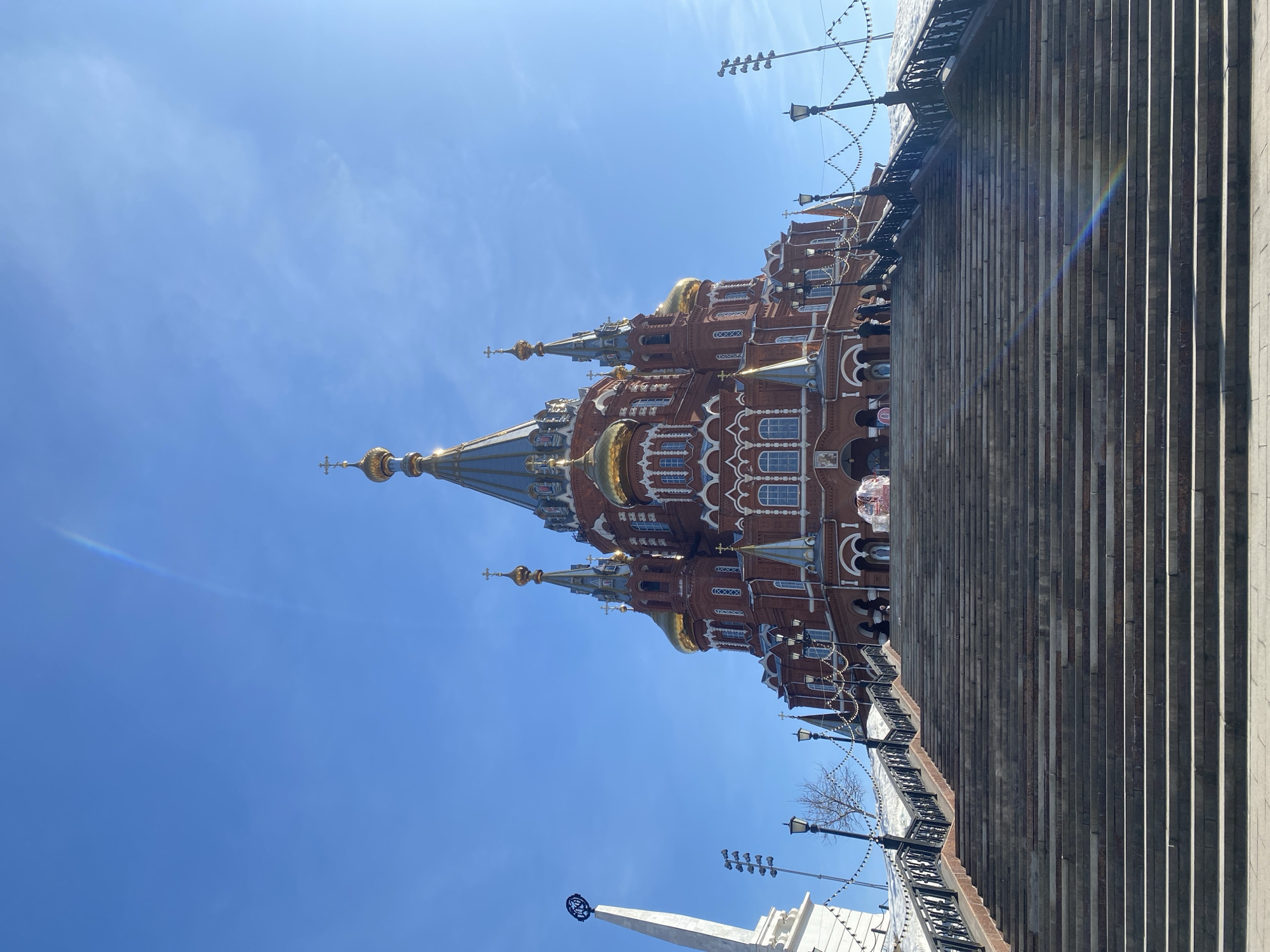 The image shows the Mikhailovsky Cathedral in Izhevsk, Russia, from a low angle, looking up. The cathedral is a red brick building with multiple golden domes and intricate white architectural details. The sky is a clear blue. To the right of the cathedral are a set of stone steps, stretching upwards. Lamp posts with hanging lights are visible along the steps. People are barely visible near the cathedral's entrance. A partial view of a white building with a spire is at the bottom of the image.