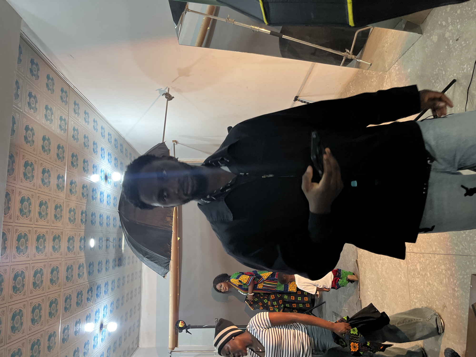 In a studio setting in Gura Top, Nigeria, a group of individuals are engaged in a photoshoot. A man in a black jacket and light blue jeans is the primary subject, holding a phone in his right hand as he looks towards the camera. To his left, an umbrella is positioned on a stand. In the background, a woman is seated, wearing a vibrantly patterned outfit. Another person, wearing a striped shirt and a beanie, is also present, holding fabric. The studio walls are decorated with a patterned tile design. Lights and photography equipment are visible throughout the scene, indicating the ongoing activity. The lighting suggests an indoor environment, likely during the day. The overall atmosphere appears to be a professional or creative collaboration.