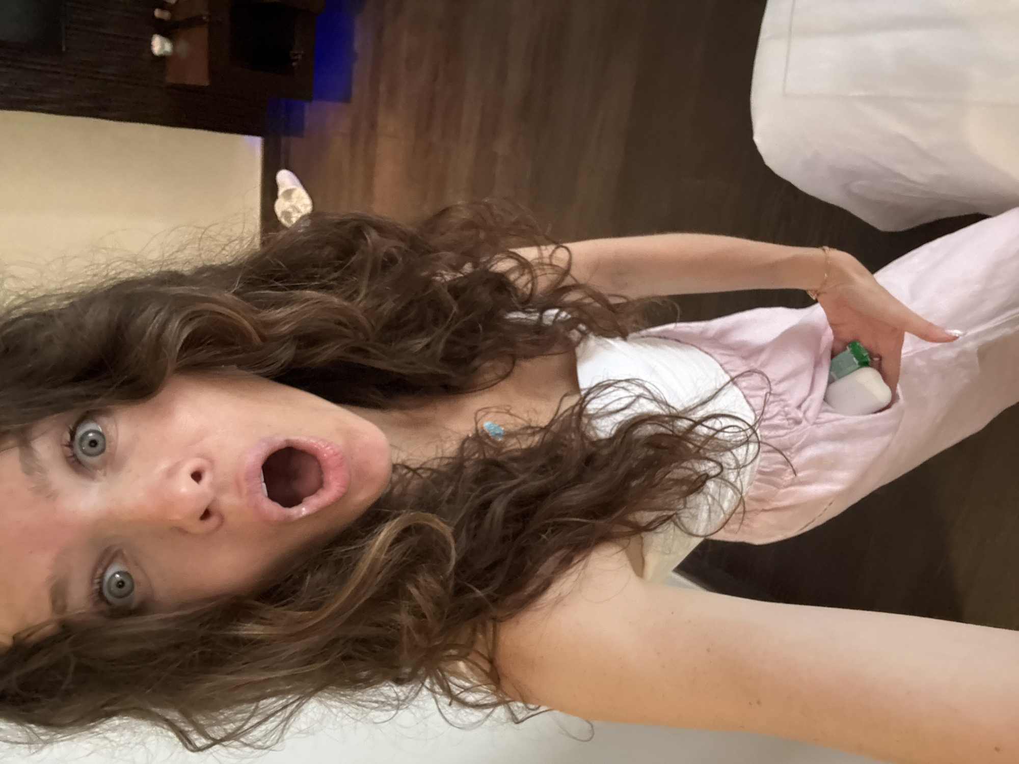 A woman with curly brown hair and wide blue eyes is looking directly at the camera with her mouth open in a surprised expression. She is wearing a white strapless top and light pink pants. Her left hand is visible, holding a small white object with a green top. The background appears to be an indoor setting with a wooden floor and a dark wooden piece of furniture. A pair of white slippers with a heart design are visible on the floor to the right of the woman. The lighting is warm and casts soft shadows.