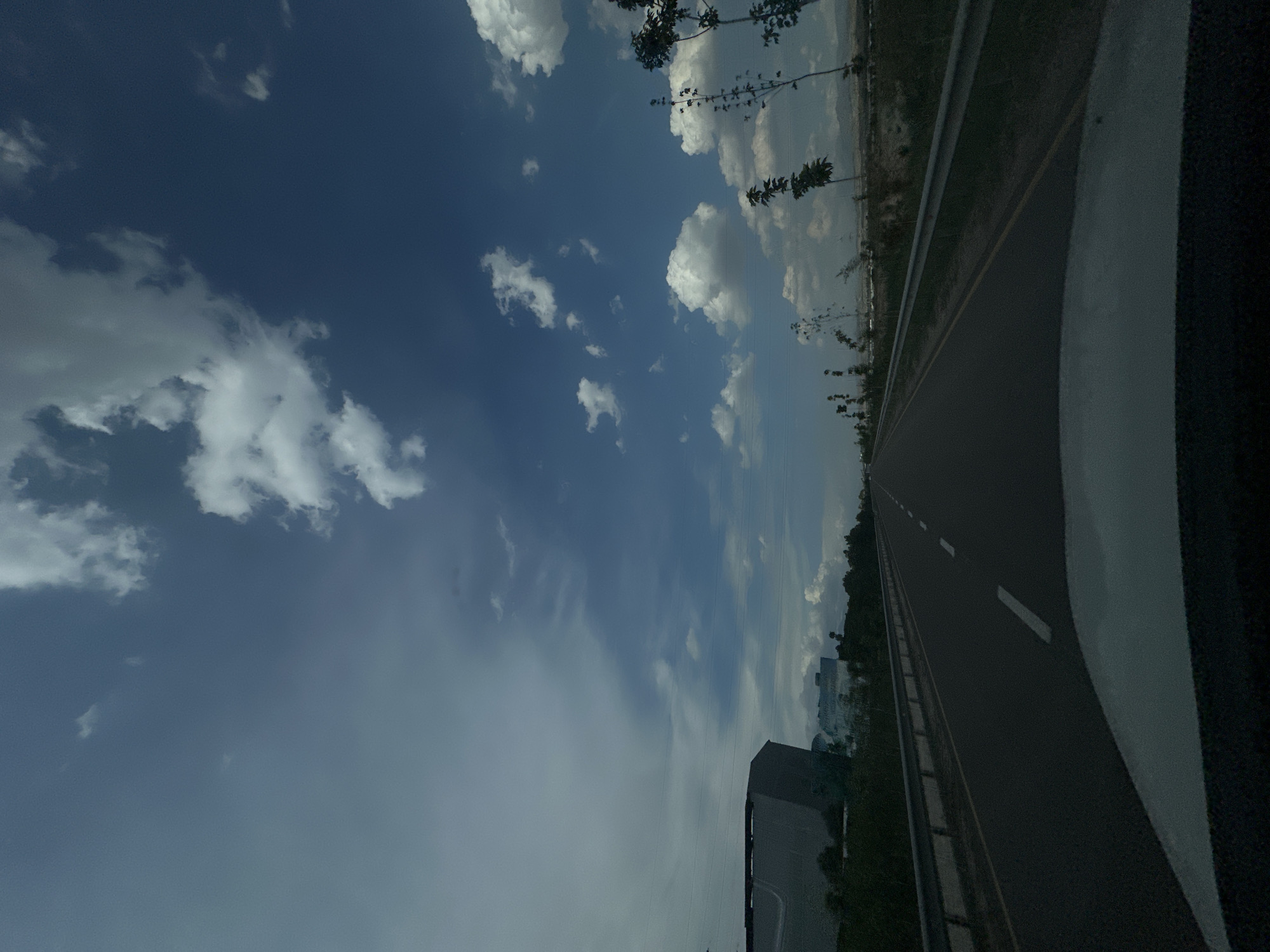 The image depicts a roadway in Nalazar, Uzbekistan, under a sky filled with scattered white cumulus clouds against a deep blue expanse. The perspective is from the driver's seat of a vehicle, looking forward along an asphalt road with white dashed lane markings. On the left side of the road, a metal guardrail runs parallel to the asphalt, bordered by a strip of yellow. Beyond the guardrail, a large, modern building with a dark, angular facade is visible, situated amongst sparse greenery. Further in the distance on the left, another, lighter-colored building can be seen. The right side of the road features a similar guardrail, followed by a dry, barren landscape with scattered young trees and shrubs. Mountains are faintly visible on the horizon in the far distance on the right. The overall impression is a highway stretch in a semi-urban or developing area, with signs of construction and infrastructure. There are no people or animals visible in the scene.