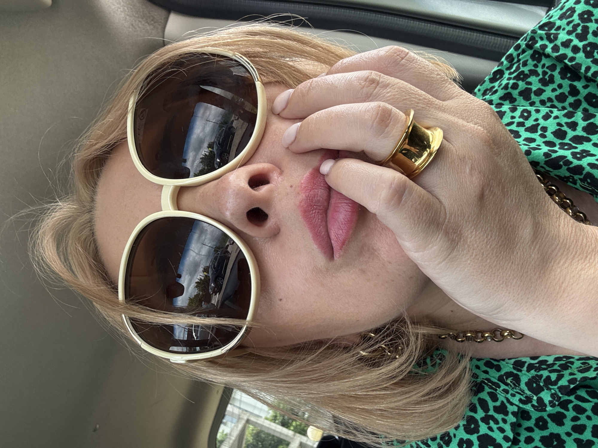 The image is a close-up, rotated photograph of a person wearing oversized sunglasses with light-colored rims. The person has blonde hair, and is wearing a bright green top with a black leopard print. A large gold ring is visible on the person's hand, which is touching their face near their lips. A gold chain necklace is also visible. The scene appears to be inside a vehicle, suggested by the interior lighting and the reflection of trees and cars in the sunglasses. The image is likely taken during the daytime, given the brightness and reflections. The person's makeup is noticeable, with pink lipstick and light-colored nail polish. There is no visible text in the image.