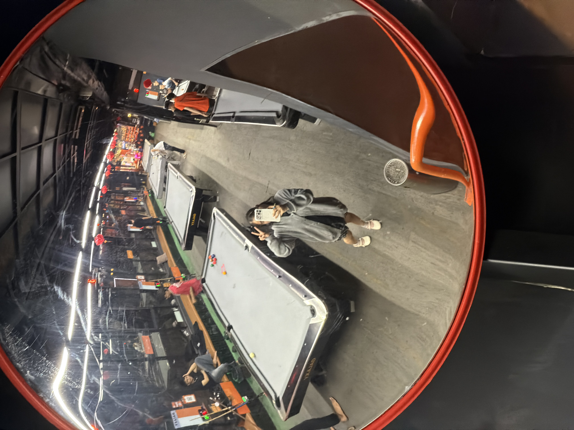 The image displays a wide, distorted reflection of an indoor billiard hall, captured in a large, circular convex mirror with a bright red frame.
The main subject is a person taking a selfie within the reflection, holding a white smartphone with one hand and making a peace sign with the other. This individual is dressed in a grey hooded sweatshirt and light-colored sandals.
The scene features multiple grey-topped pool or billiard tables, some with colorful balls scattered across them. Several other people are visible in the background, appearing to be playing or socializing. One person in an orange shirt is seen near a table on the right side of the reflection, while another is seated near a table on the left. The hall is illuminated by artificial lighting, creating bright, streaky reflections across the surfaces. Red spherical decorations, possibly lanterns, are visible hanging within the space.
Notable details include a circular drainage grate on the floor near the mirror, an orange, curved support structure, and the industrial-style concrete floor. The convex mirror distorts proportions, offering a broad, panoramic view of the establishment. Visible text "YRAJAD" appears in reverse on the side of the pool tables, likely indicating the name "DAJARY." The setting is an active, casual indoor environment, consistent with a recreational venue in Đà Nẵng, Vietnam.