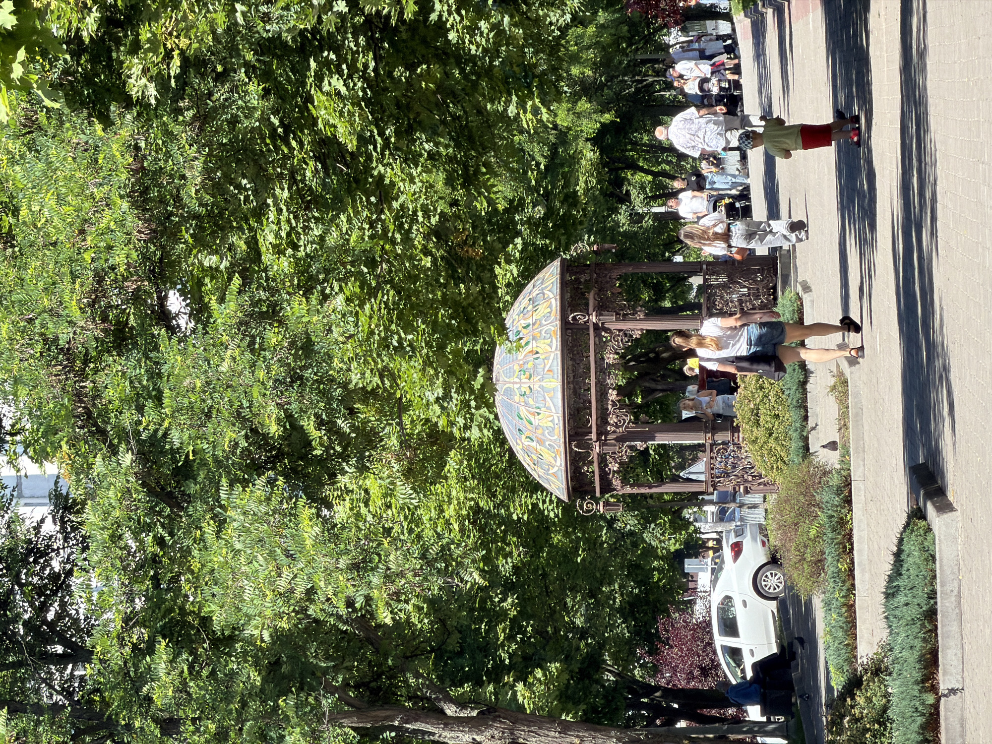 The image depicts a sunny day scene in a park in Dnipro, Ukraine. In the foreground, a paved walkway is lined with green bushes and trees. Several people are walking along the path, including a woman in a white t-shirt and denim shorts, a child in a green shirt and red shorts riding a scooter, and a man in a patterned shirt. Further down the path, more individuals and a stroller are visible. To the left, a white car is parked near a bench where a person is seated. In the background, a decorative gazebo with a stained-glass dome stands among the lush green foliage. The overall atmosphere suggests a leisurely public space.