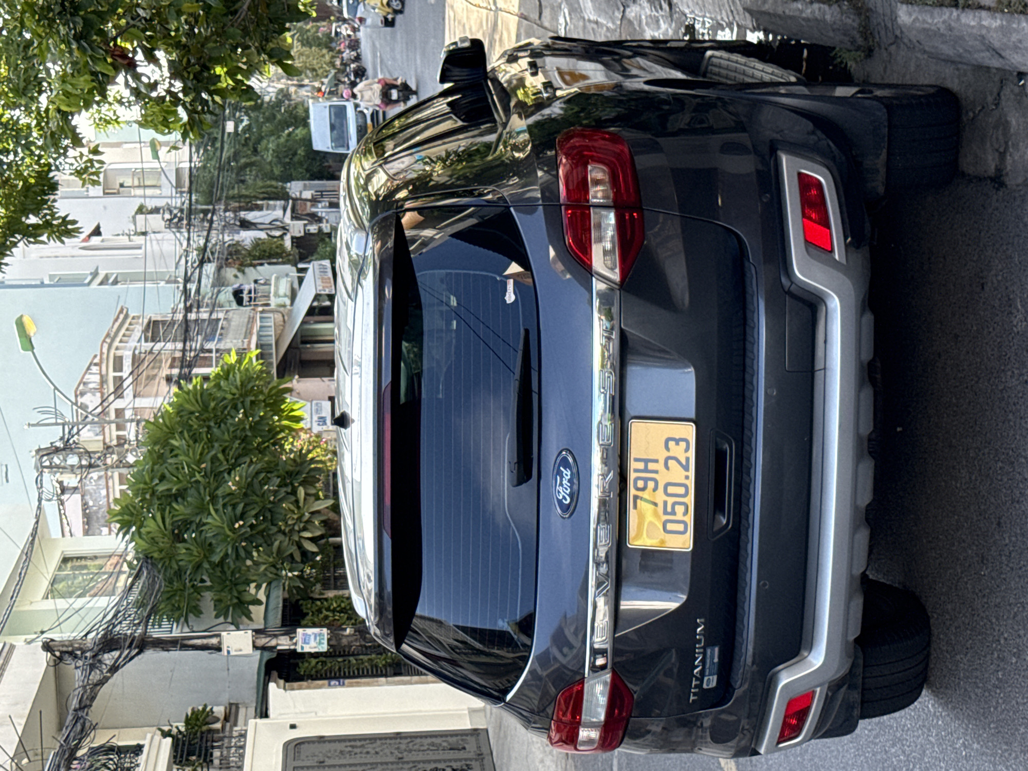 The image shows the rear of a dark gray Ford Everest SUV parked on the side of a street in Nha Trang, Vietnam. The license plate reads "79H 050.23". The car has "TITANIUM" badging on the lower left of the tailgate. The street is lined with buildings, power lines, and trees. In the background, other vehicles, including a white van and several motorcycles, are visible on the street, along with a few pedestrians. The sun appears to be shining, casting shadows.