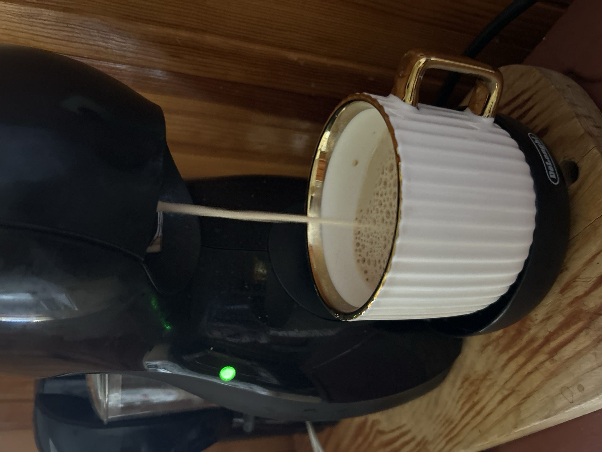 An overhead shot shows a close-up of a black coffee maker dispensing a light brown liquid into a white mug with a gold rim and handle. The mug is sitting on a black saucer, which is on a wooden surface. The coffee maker has a green light on its side. The background is a wooden wall. The image does not provide any information about the location in the city of Basarabeasca, Moldova.