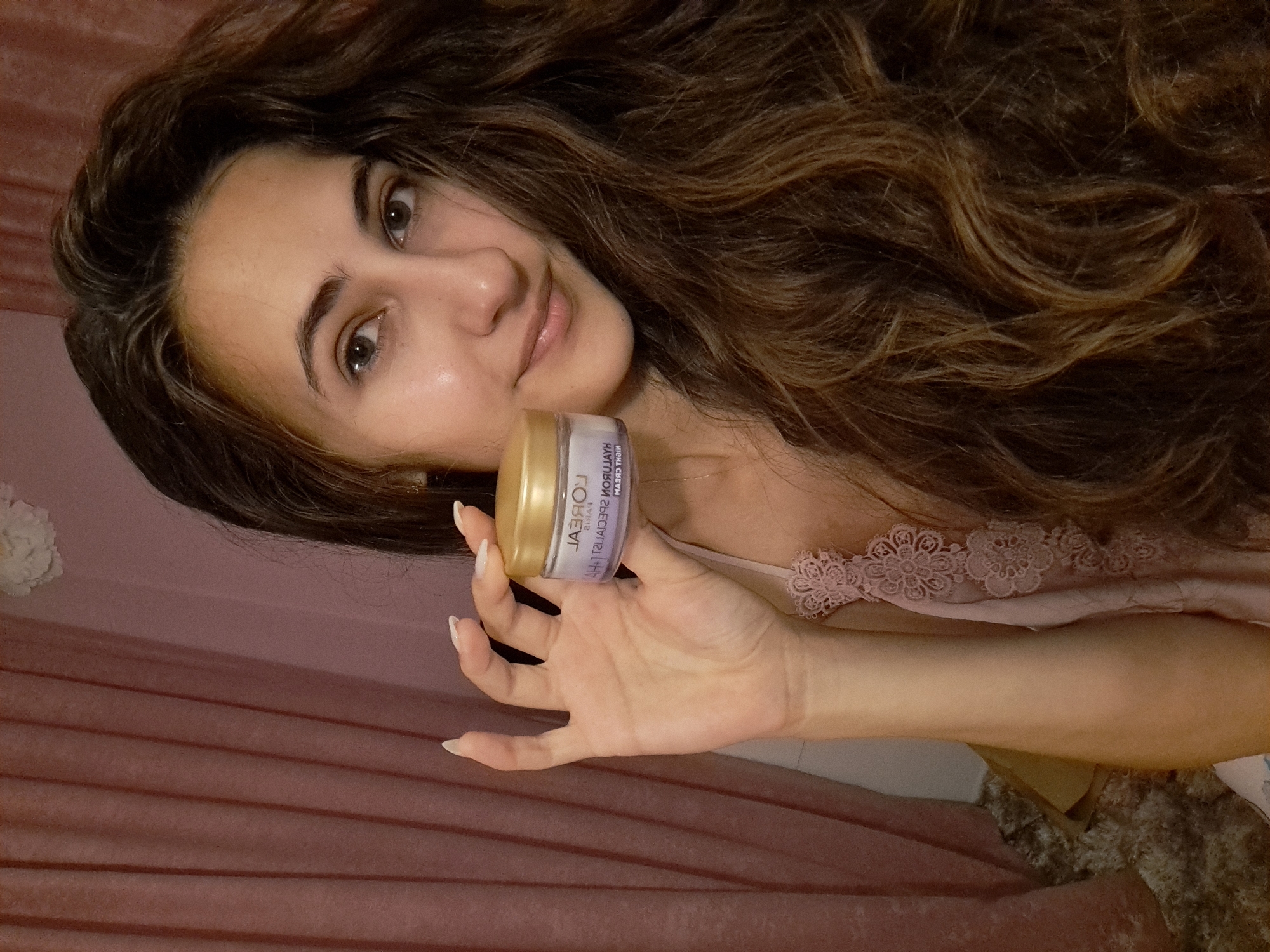 A young woman with long, wavy brown hair holds a small, round jar of L'Oréal Paris Hyaluron Specialist Night Cream. She is wearing a light pink, lacy camisole. The background consists of dusty rose-colored curtains and a textured rug. The lighting is soft and diffused. The woman is looking directly at the camera with a slight smile. The image appears to be taken indoors, possibly in a bedroom.