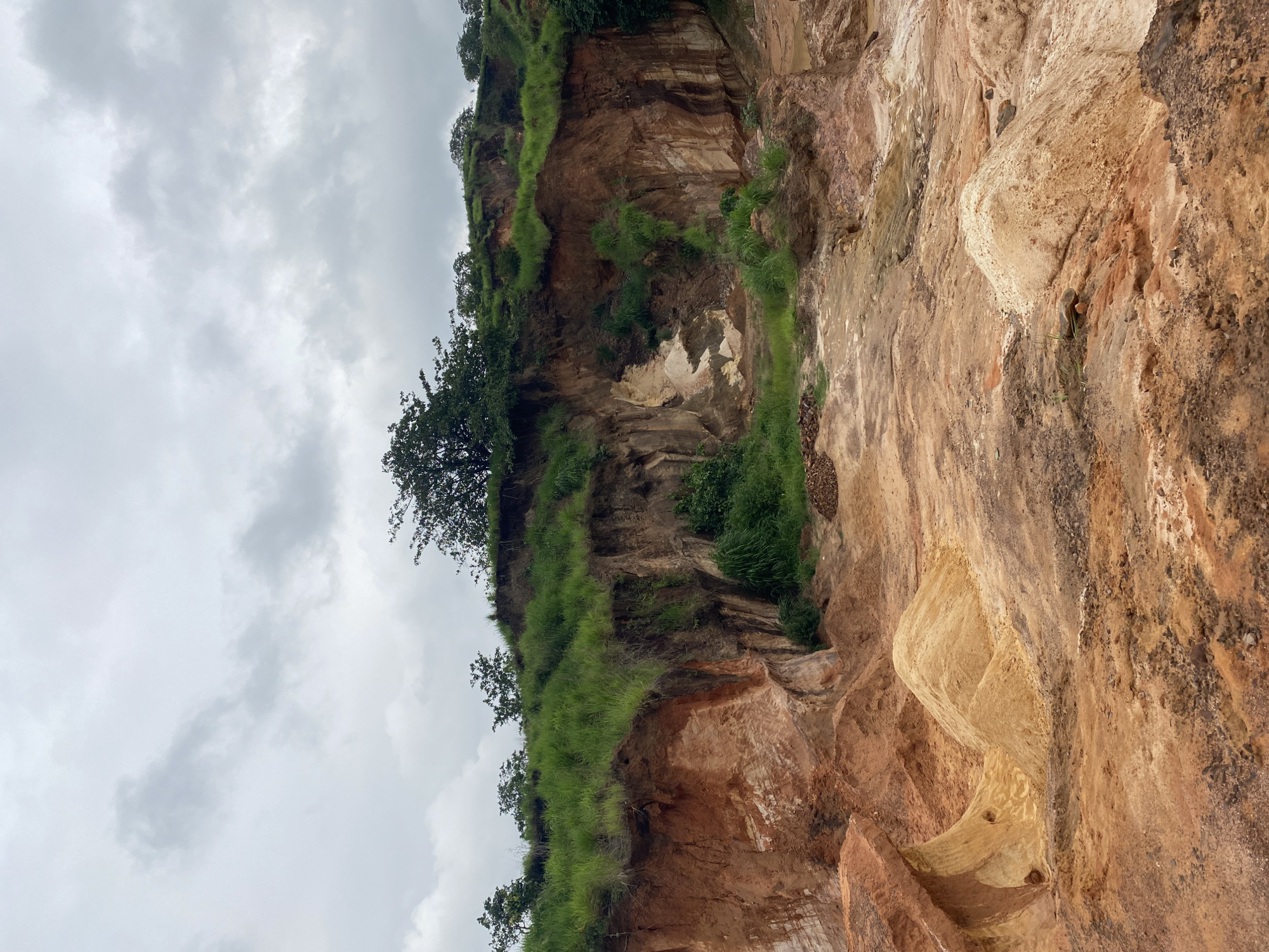 The image depicts a quarry site in Achara, Nigeria. The landscape is characterized by eroded earth with exposed layers of reddish-brown and pale yellow soil. Vegetation, primarily green grass and scattered trees, grows along the rim of the quarry and in some crevices. The sky is overcast with grey clouds. In the foreground, mounds of excavated soil are visible. The scene appears to be devoid of human presence or recent activity.