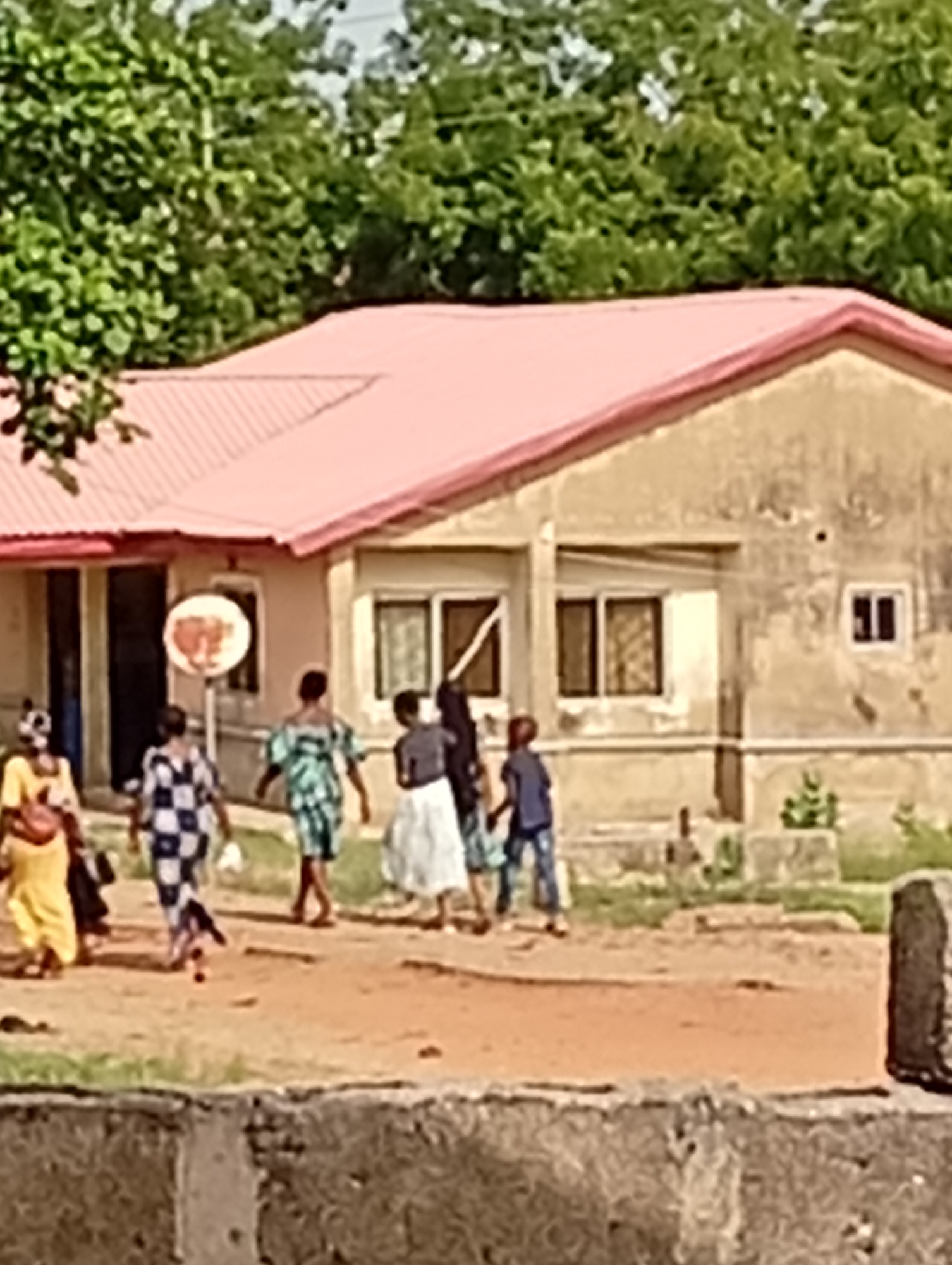 The image shows a street scene in Adankolo, Nigeria. A single-story building with a pink roof is visible in the background. Several individuals are walking along the unpaved road in front of the building. A woman in a yellow dress walks to the left. Another woman in a blue and white checkered garment walks next to the woman in the yellow dress. Next to the checkered dress there is a woman wearing a green and blue dress. Then there is a women with a gray shirt and white skirt. Following her is a small boy in a blue shirt and blue jeans. A circular sign is visible near the entrance to the building. The foreground shows a concrete structure. Trees are visible in the distance behind the building.