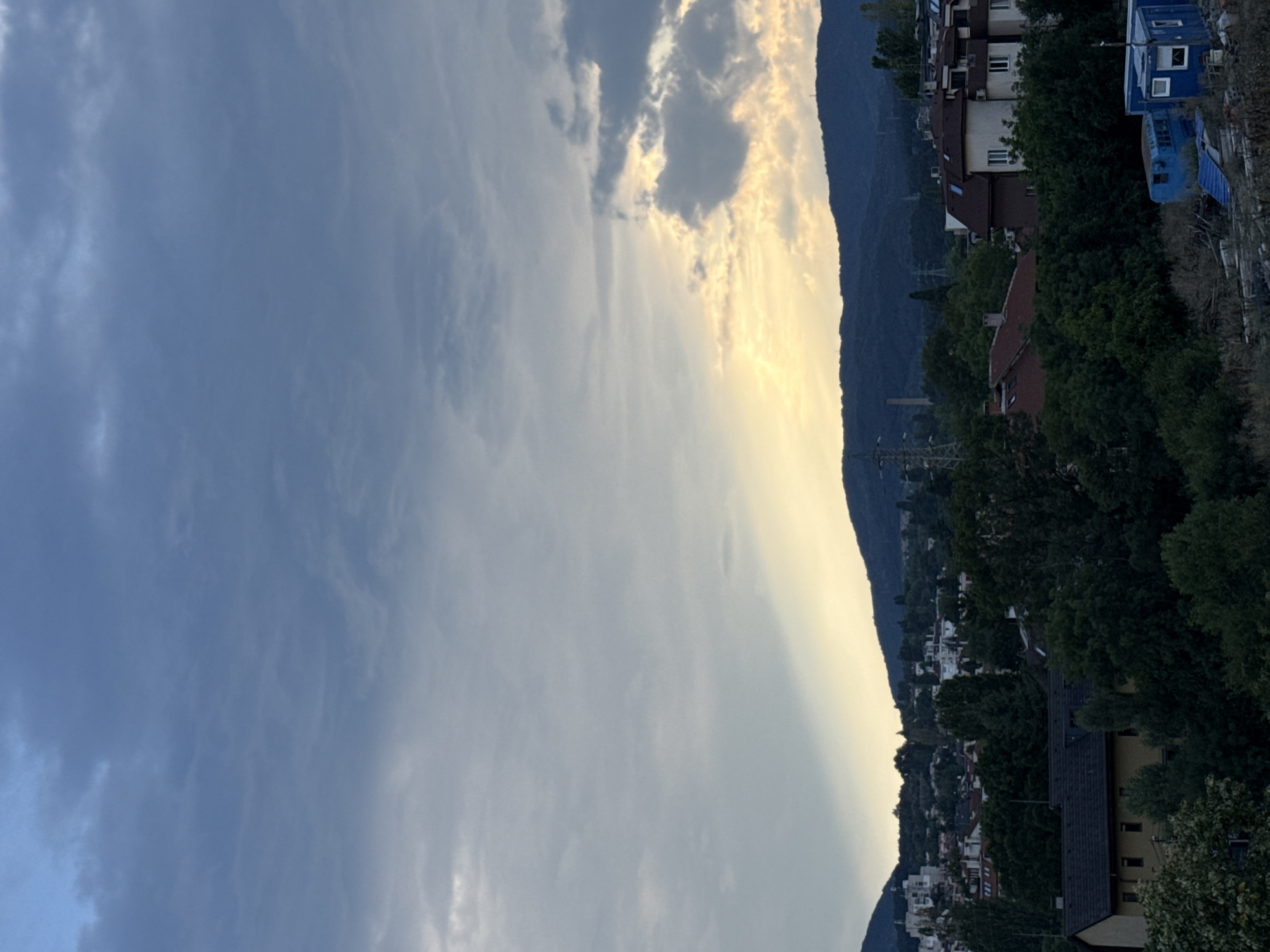 The image captures a twilight scene over a suburban area in Sofia, Bulgaria, likely in the western or southwestern part of the city given the mountainous backdrop. The sky is a mix of dramatic grey clouds with shafts of golden light breaking through, suggesting the sun is either setting or has just risen behind the clouds and hills.
In the foreground, a collection of trees and vegetation obscures parts of the view. A light-colored building with a dark-tiled roof is partially visible on the left, surrounded by dense greenery. Further into the midground, there are clusters of houses and buildings, some with red-tiled roofs and others with lighter facades, typical of residential architecture in Sofia. Power lines and pylons are visible among the trees, indicating the presence of infrastructure.
In the far distance, dark, rolling hills form the horizon, with a few faint structures, possibly towers or antennas, visible against the skyline. In the immediate foreground on the right, a cluttered area with debris is present, featuring two blue modular structures, one appearing to be a trailer or shed with windows, and another smaller blue container-like unit. The overall atmosphere is serene but with a touch of urban sprawl meeting natural landscape. No people are visible in the image.
