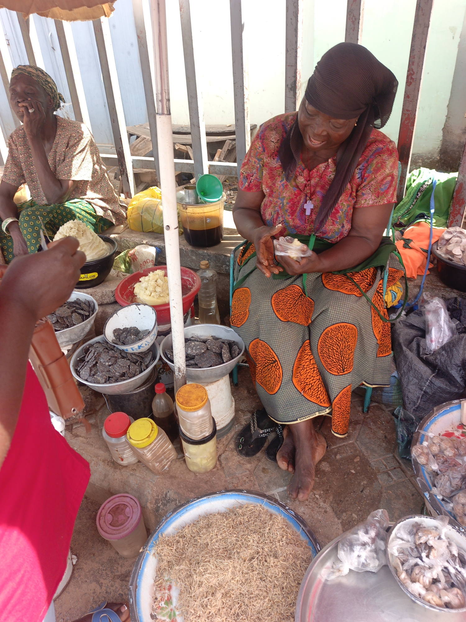 The image captures a market scene in Akwanga, Nigeria. An elderly woman wearing a patterned top, a green and orange skirt, and a dark headwrap sits on a blue stool, smiling as she wraps a small portion of white butter-like substance in plastic. Bowls surrounding her contain what appear to be dried turtle shells, shea butter, and unidentified dried goods. Another woman sits to the left, dressed in a patterned top and green skirt, with bracelets on her wrist. Jars, plastic containers, and buckets containing various ingredients are arranged nearby. A red shirt is visible in the left corner. The setting is shaded by an umbrella and a structure with metal bars.
