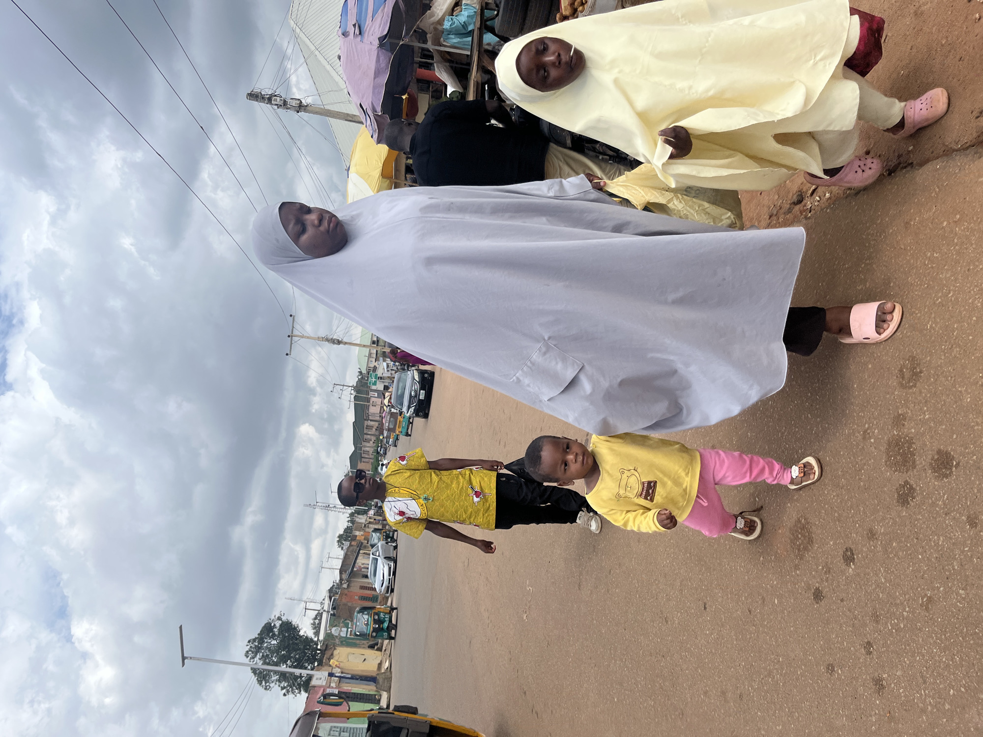 An overcast day in Jos, Nigeria, is captured in this image of a street scene. In the foreground, a young woman in a light gray hijab and a young child in a yellow hijab are walking. The woman holds a plastic bag. Next to them, a young boy in a yellow shirt with a cartoon cat print and pink pants walks barefoot. Further back, a boy in a yellow patterned shirt and sunglasses walks while holding a black plastic bag. In the background, the street is lined with buildings and market stalls. Several vehicles, including cars and a three-wheeled auto-rickshaw, are visible on the road. The ground is asphalt and reddish-brown.