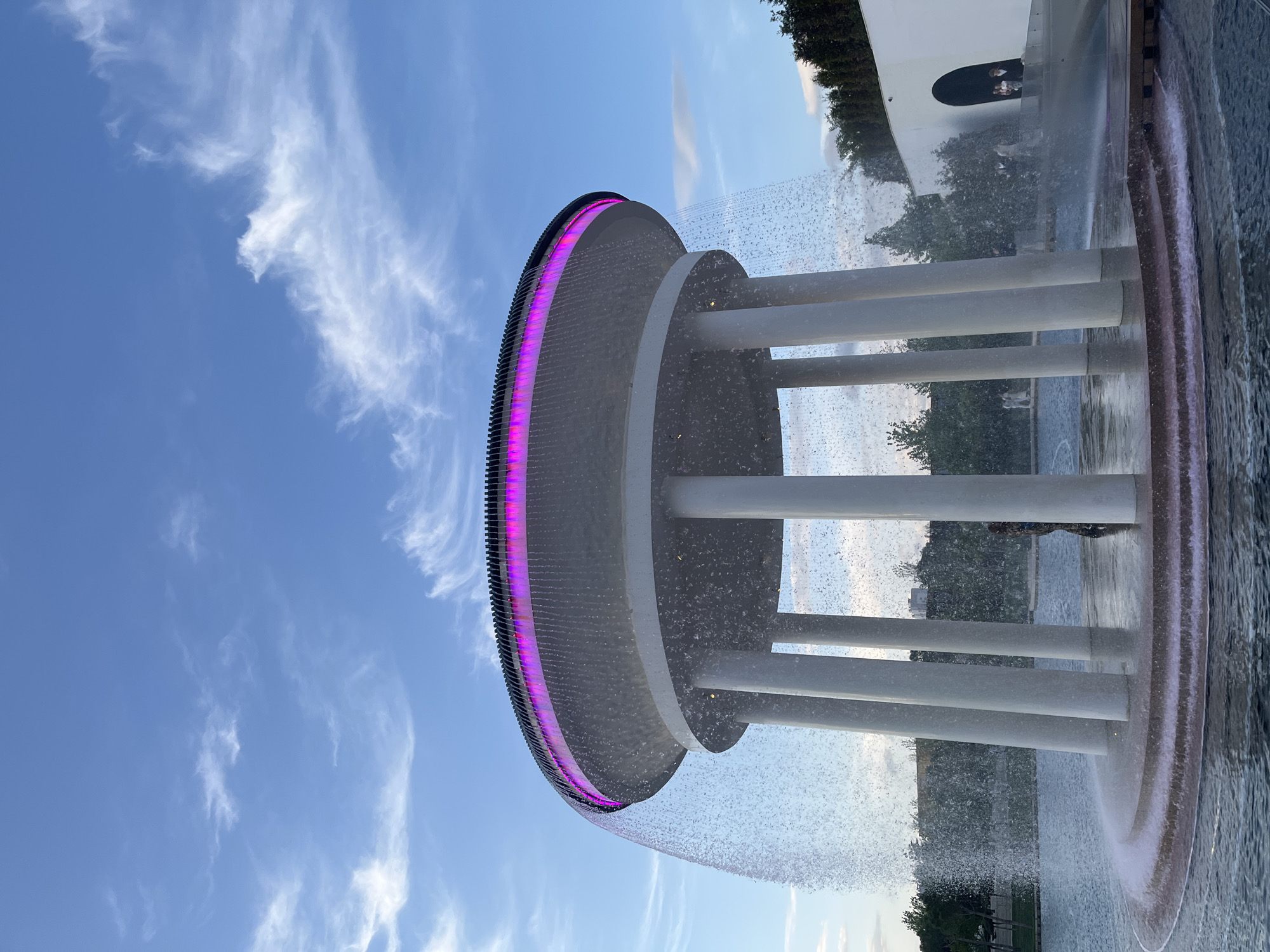 The image depicts a modern architectural structure with a water feature, set against a clear blue sky dotted with wispy clouds. The structure is circular with a pink illuminated rim and water cascading from the edge. White cylindrical supports are visible. The scene includes a body of water at the base of the structure, with reflections. In the background, there are trees and another building. The location is the Krasnodar Musical Fountain, a part of the Krasnodar Park, in Krasnodar, Russia.
