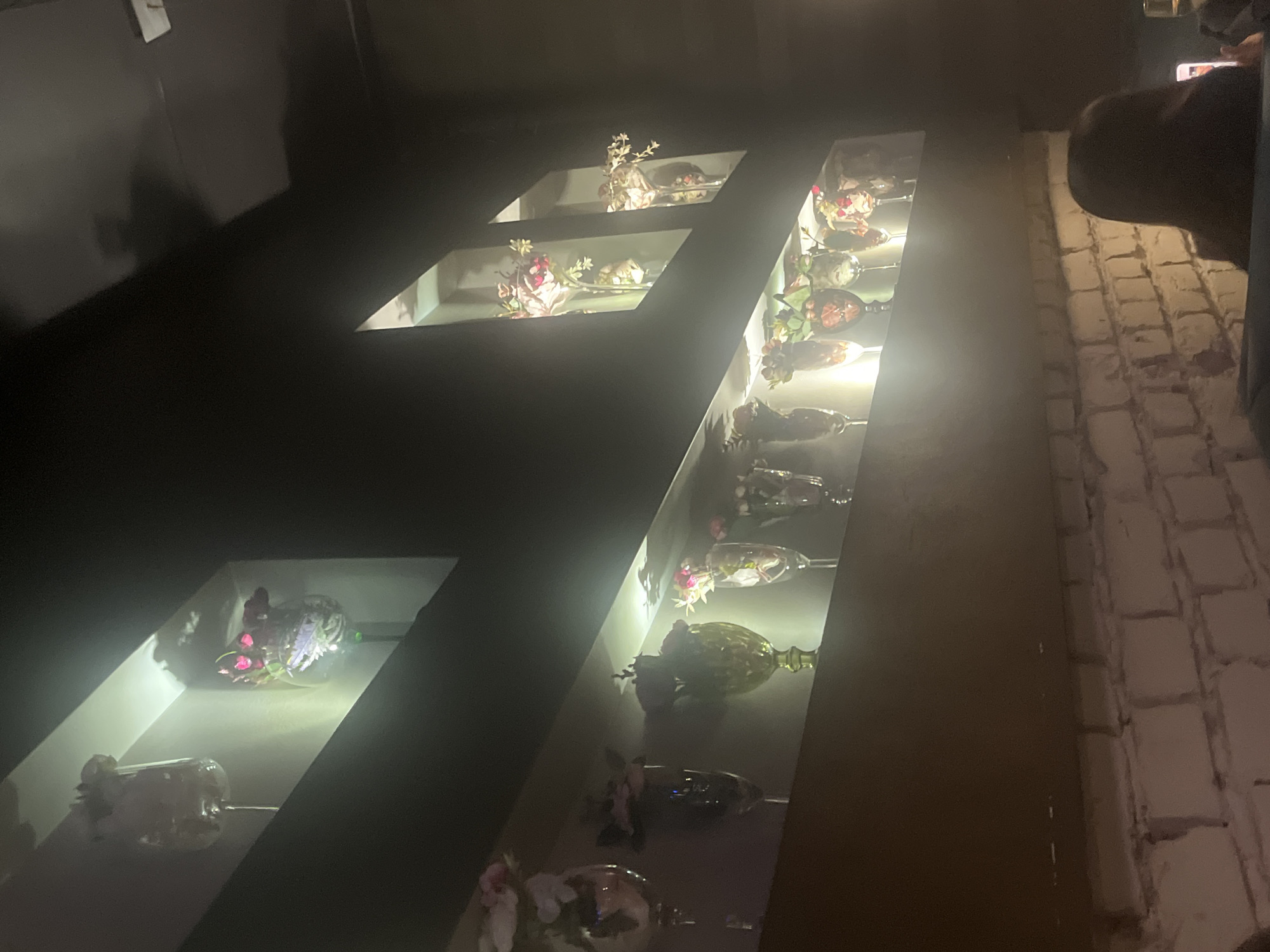 A shelf of glasses and flowers on a dark background next to a brick wall. It is possibly in a restaurant in Plovdiv, Bulgaria. A person is taking a photo of the shelf.