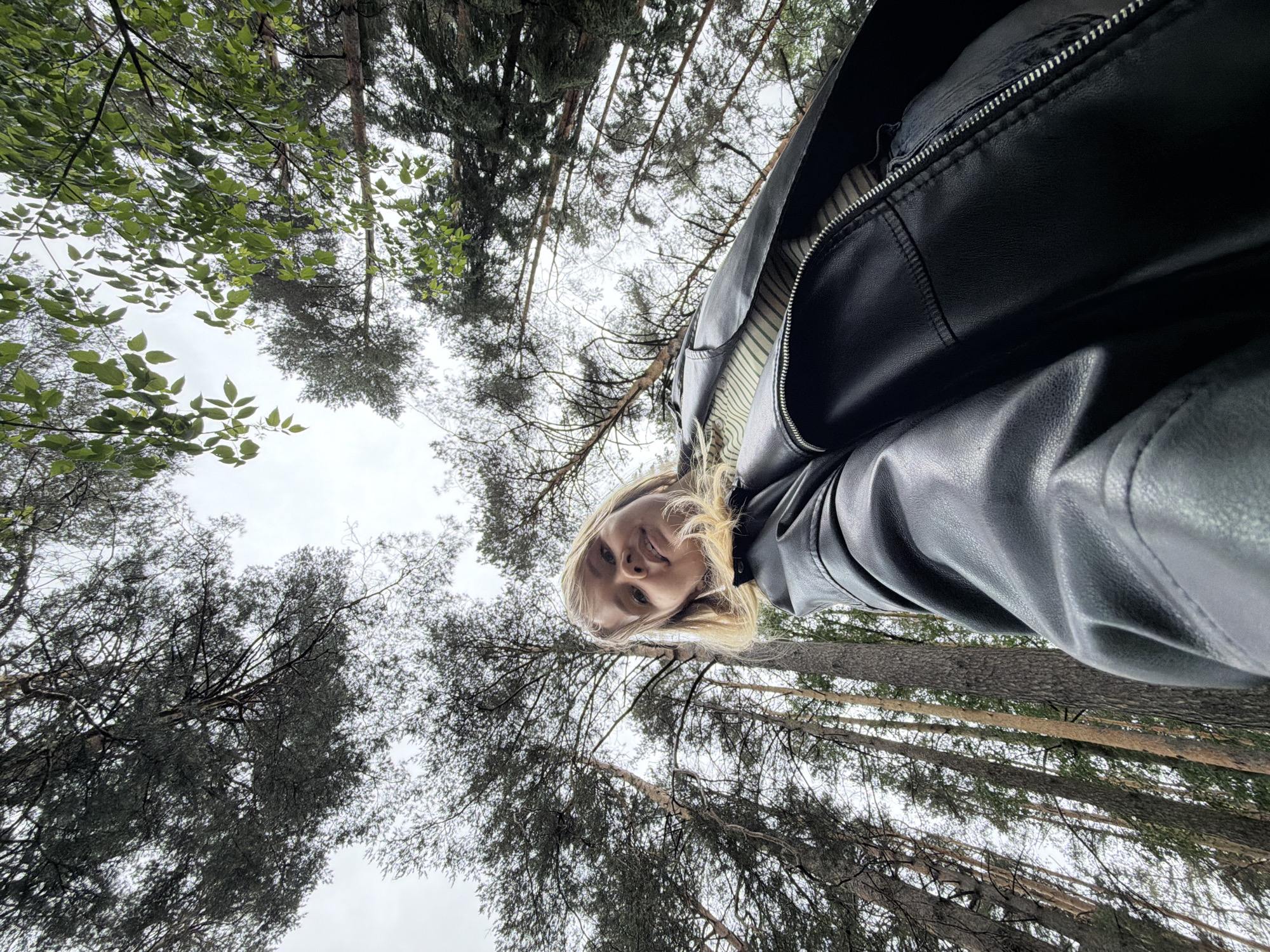 The image is an upward-facing selfie taken in a wooded area in Chaikovsky, Russia. A woman with blonde hair and wearing a black leather jacket is visible in the upper center of the frame. Tall trees surround her, with the sky visible through the branches. The trees are mostly coniferous, with some deciduous trees also present. The sky is overcast.